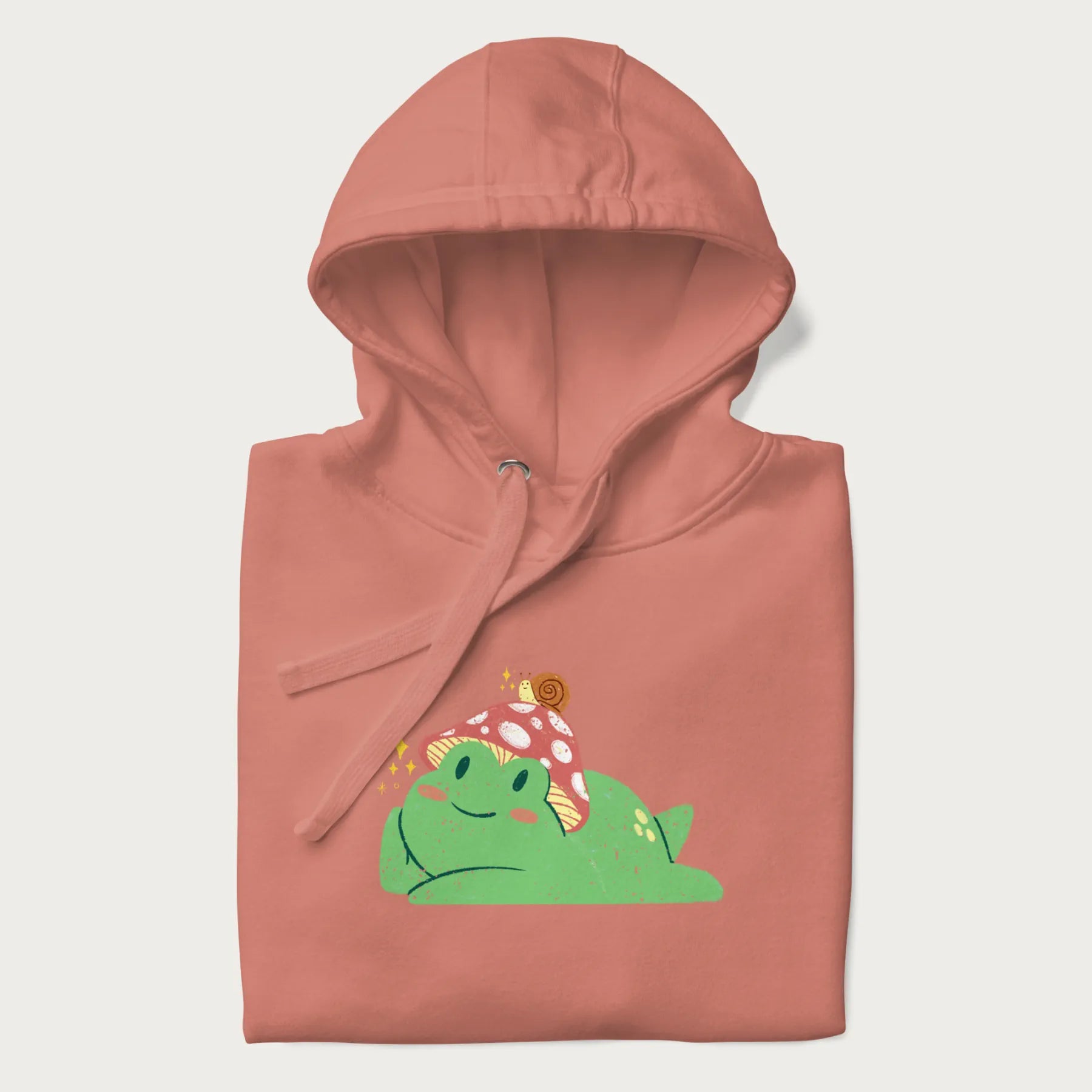 Folded light pink hoodie with a cottagecore design of a green frog wearing a mushroom cap and a snail on top.