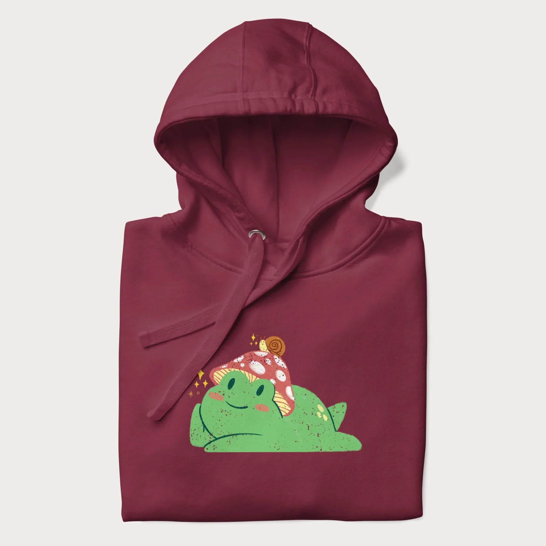 Folded maroon hoodie with a cottagecore design of a green frog wearing a mushroom cap and a snail on top.