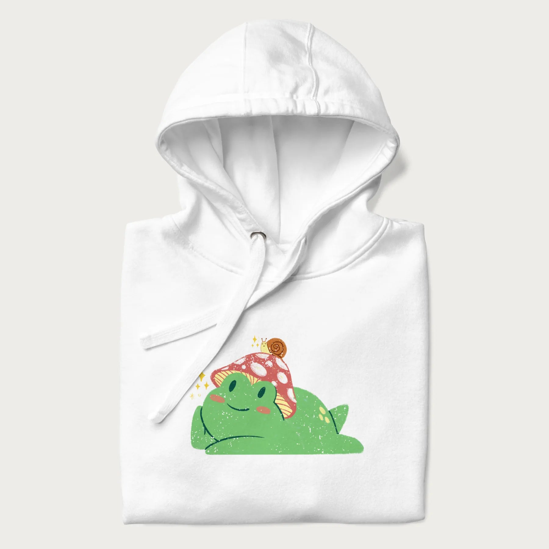 Folded white hoodie with a cottagecore design of a green frog wearing a mushroom cap and a snail on top.