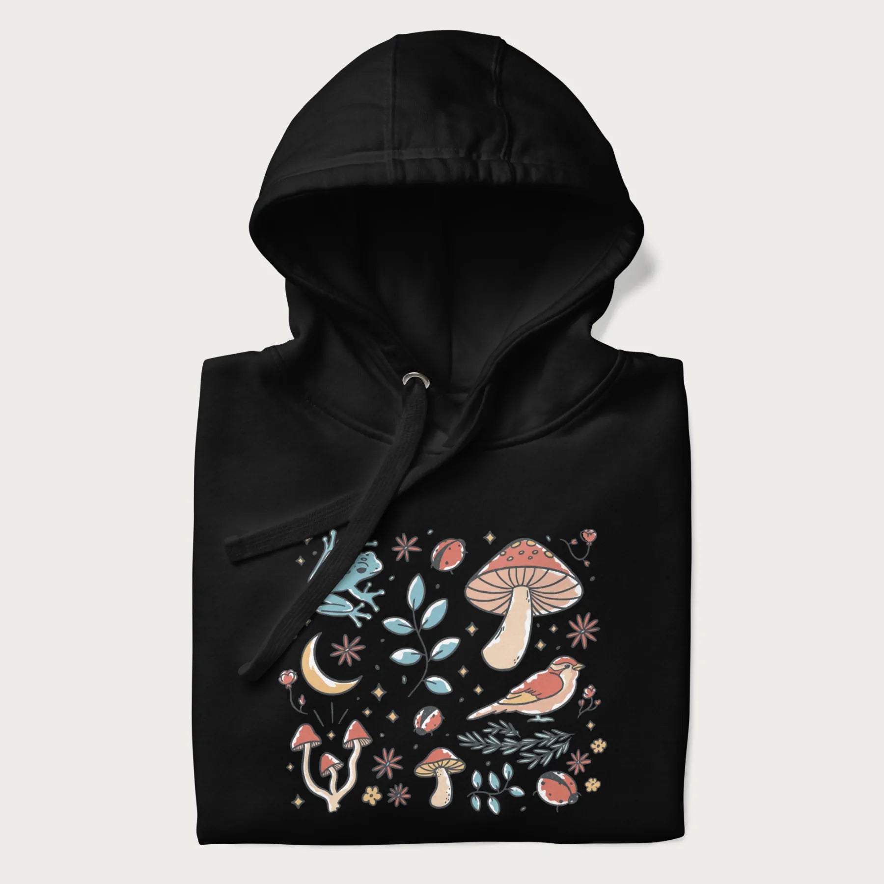 Folded black hoodie with a graphic of a blue frog, mushrooms, a bird, ladybugs, flowers, leaves, and a crescent moon.