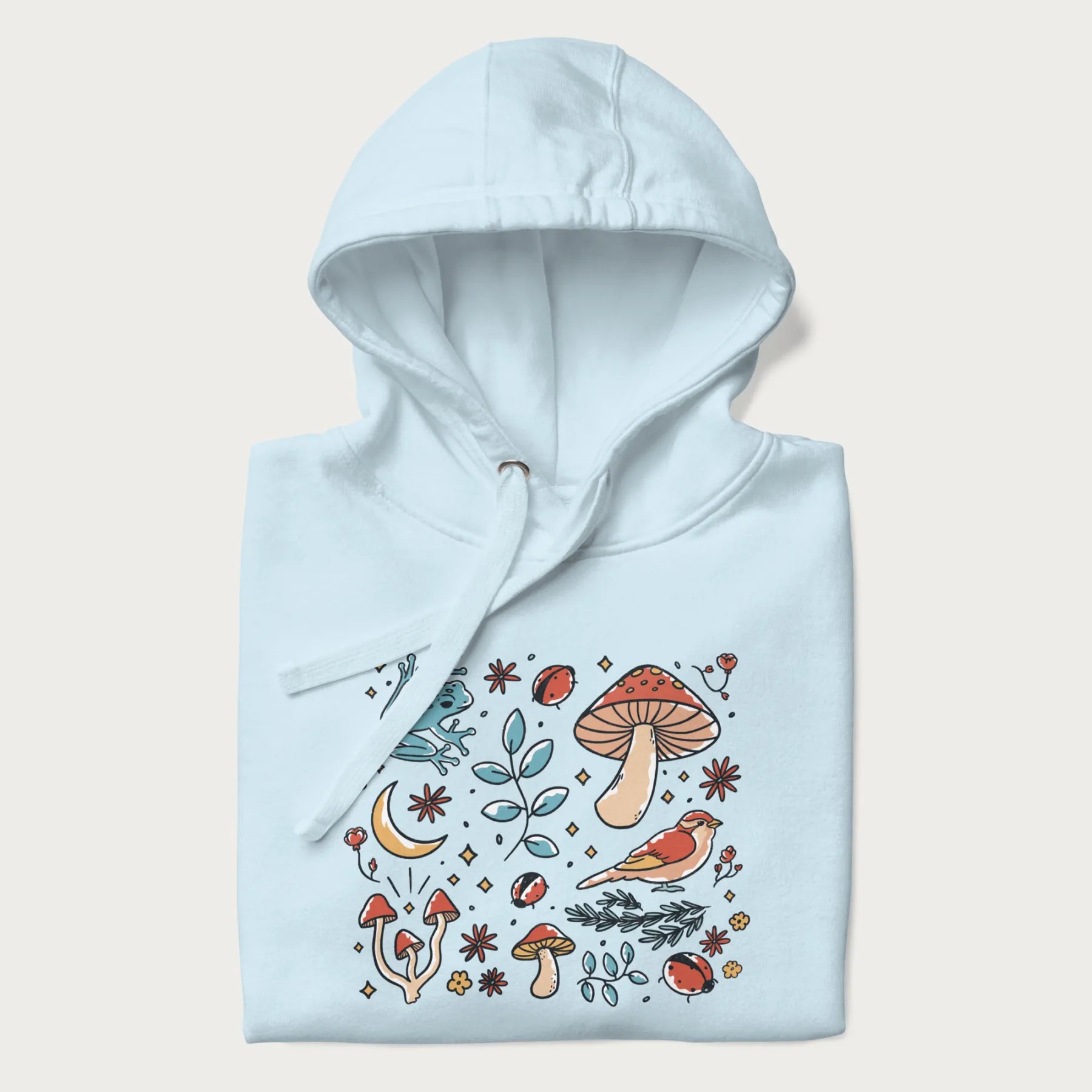 Folded light blue hoodie with a graphic of a blue frog, mushrooms, a bird, ladybugs, flowers, leaves, and a crescent moon.