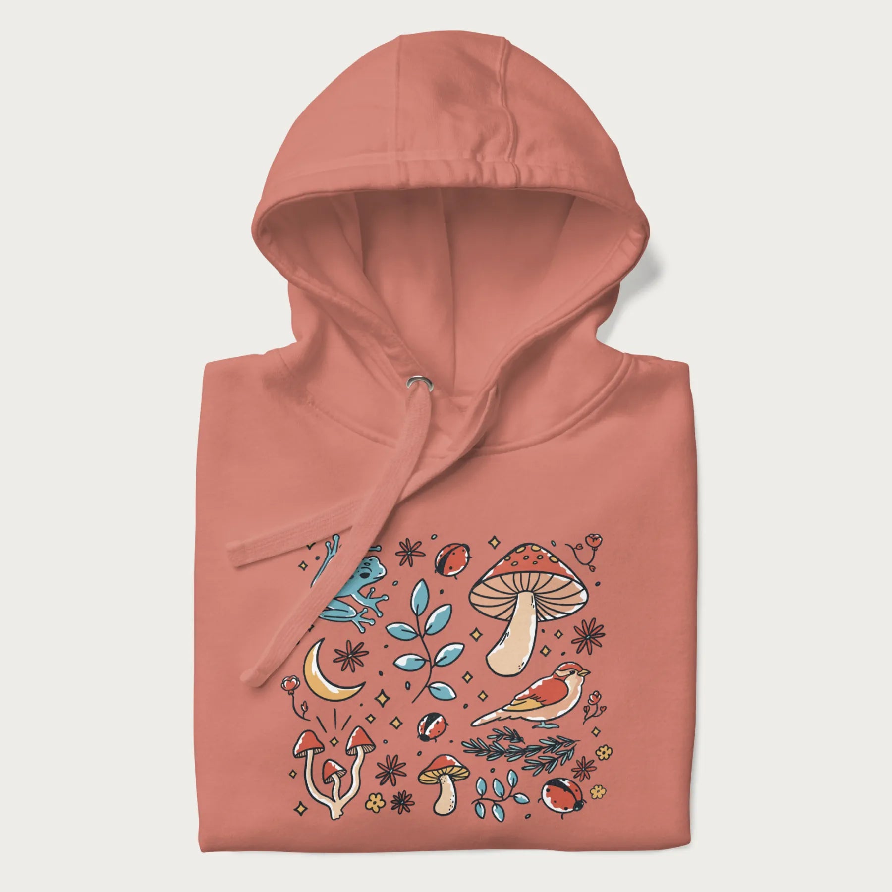 Folded light pink hoodie with a graphic of a blue frog, mushrooms, a bird, ladybugs, flowers, leaves, and a crescent moon.