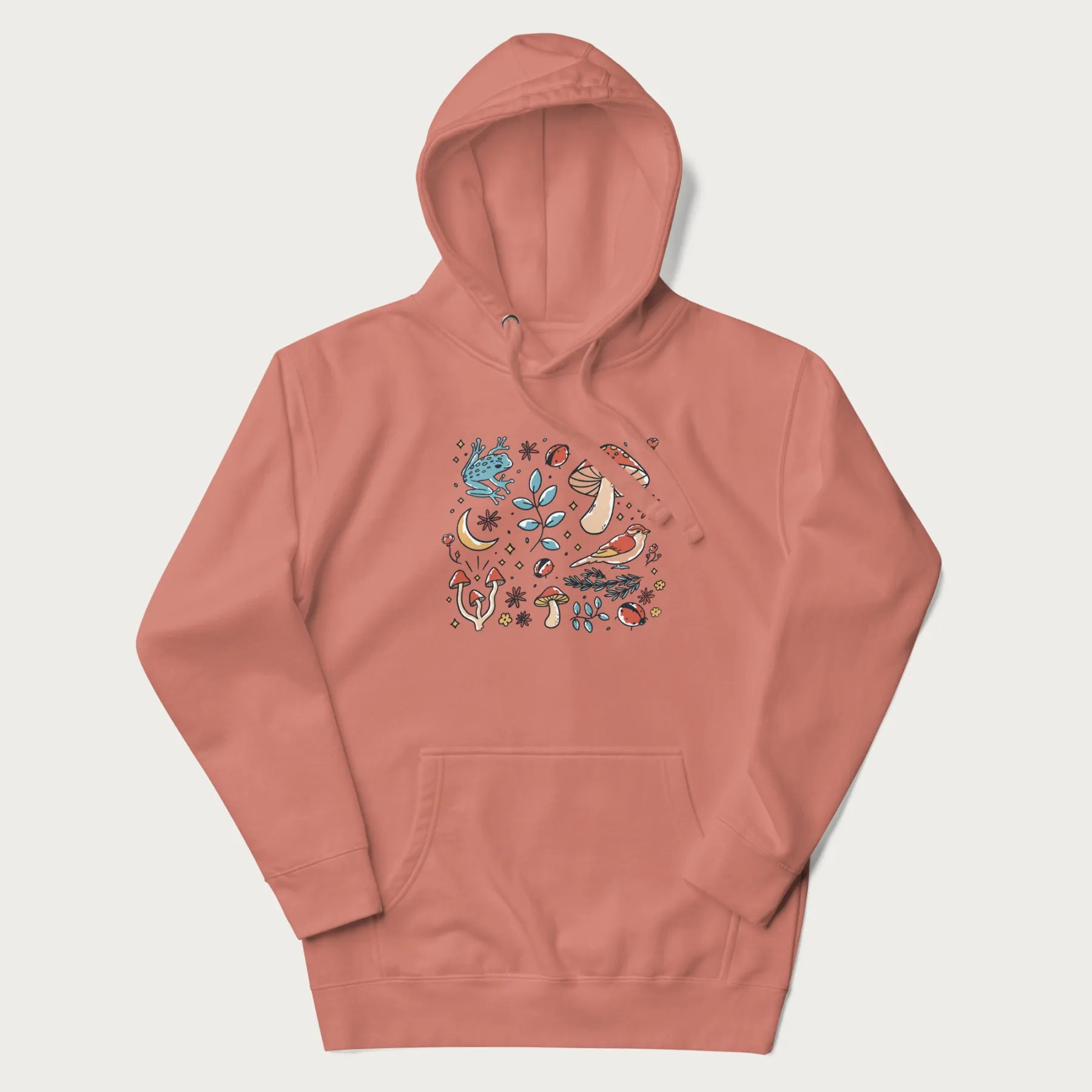 Light pink hoodie with a graphic of a blue frog, mushrooms, a bird, ladybugs, flowers, leaves, and a crescent moon.