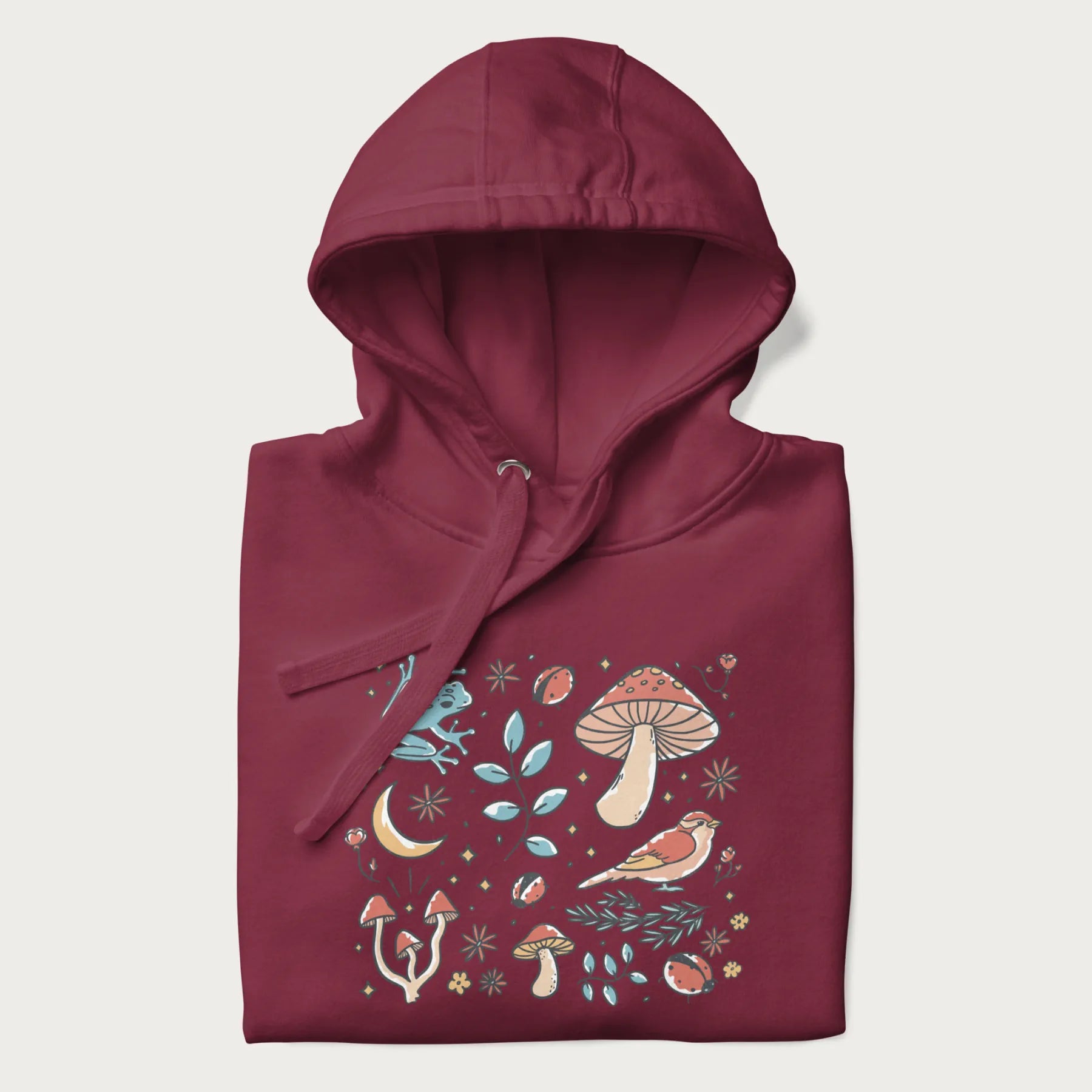 Folded maroon hoodie with a graphic of a blue frog, mushrooms, a bird, ladybugs, flowers, leaves, and a crescent moon.