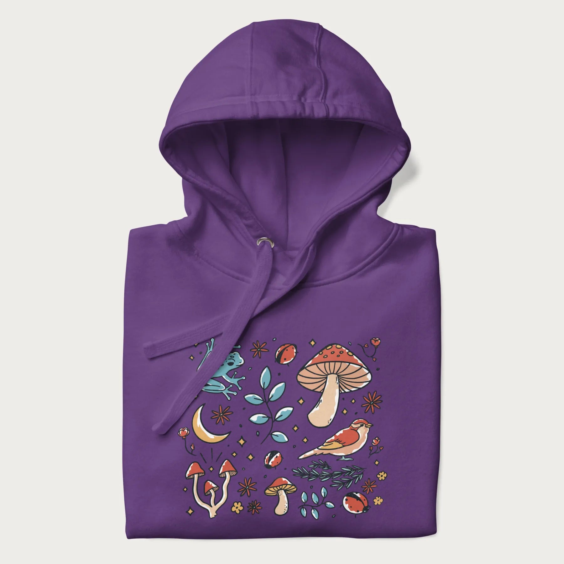 Folded purple hoodie with a graphic of a blue frog, mushrooms, a bird, ladybugs, flowers, leaves, and a crescent moon.