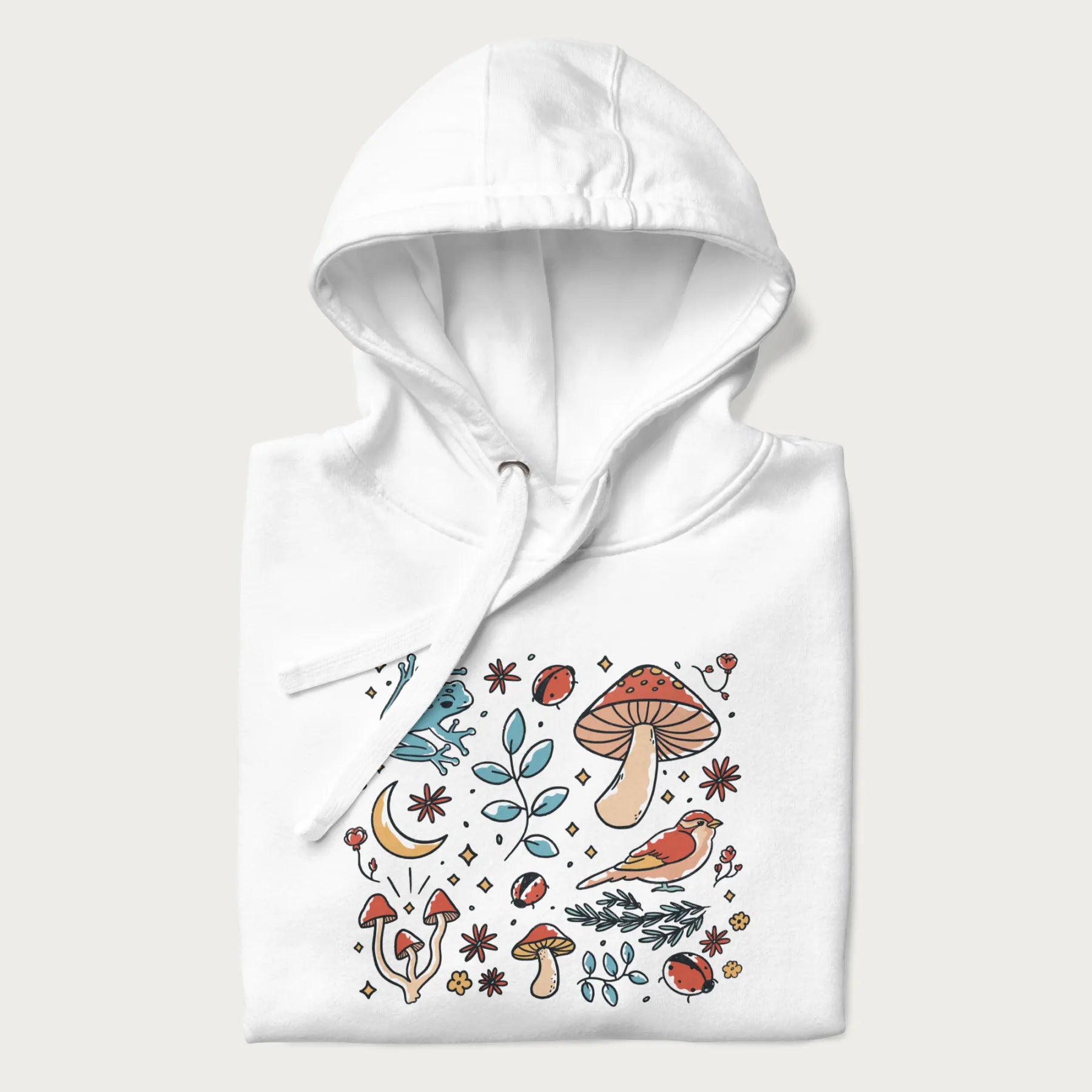 Folded white hoodie with a graphic of a blue frog, mushrooms, a bird, ladybugs, flowers, leaves, and a crescent moon.
