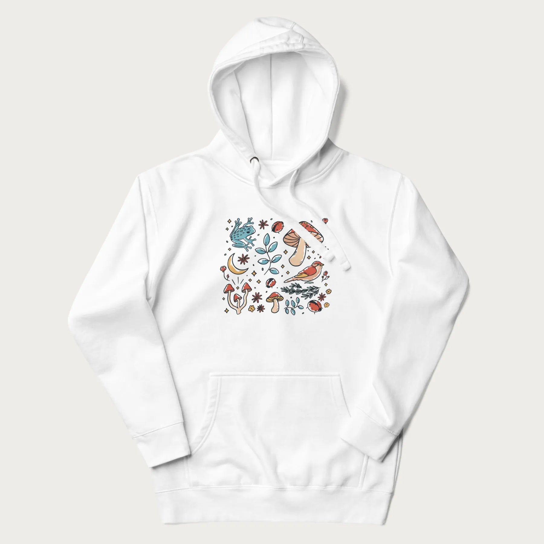 White hoodie with a graphic of a blue frog, mushrooms, a bird, ladybugs, flowers, leaves, and a crescent moon.