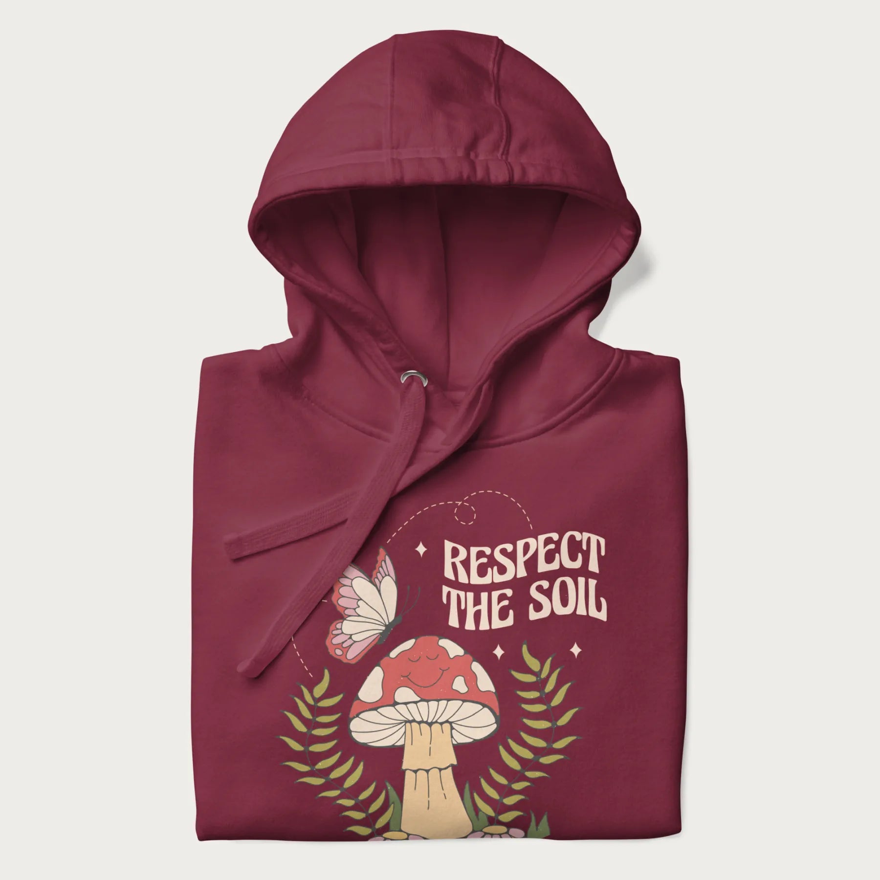 Folded maroon hoodie with mushroom and butterfly design, with the text 'Respect the Soil' above it.