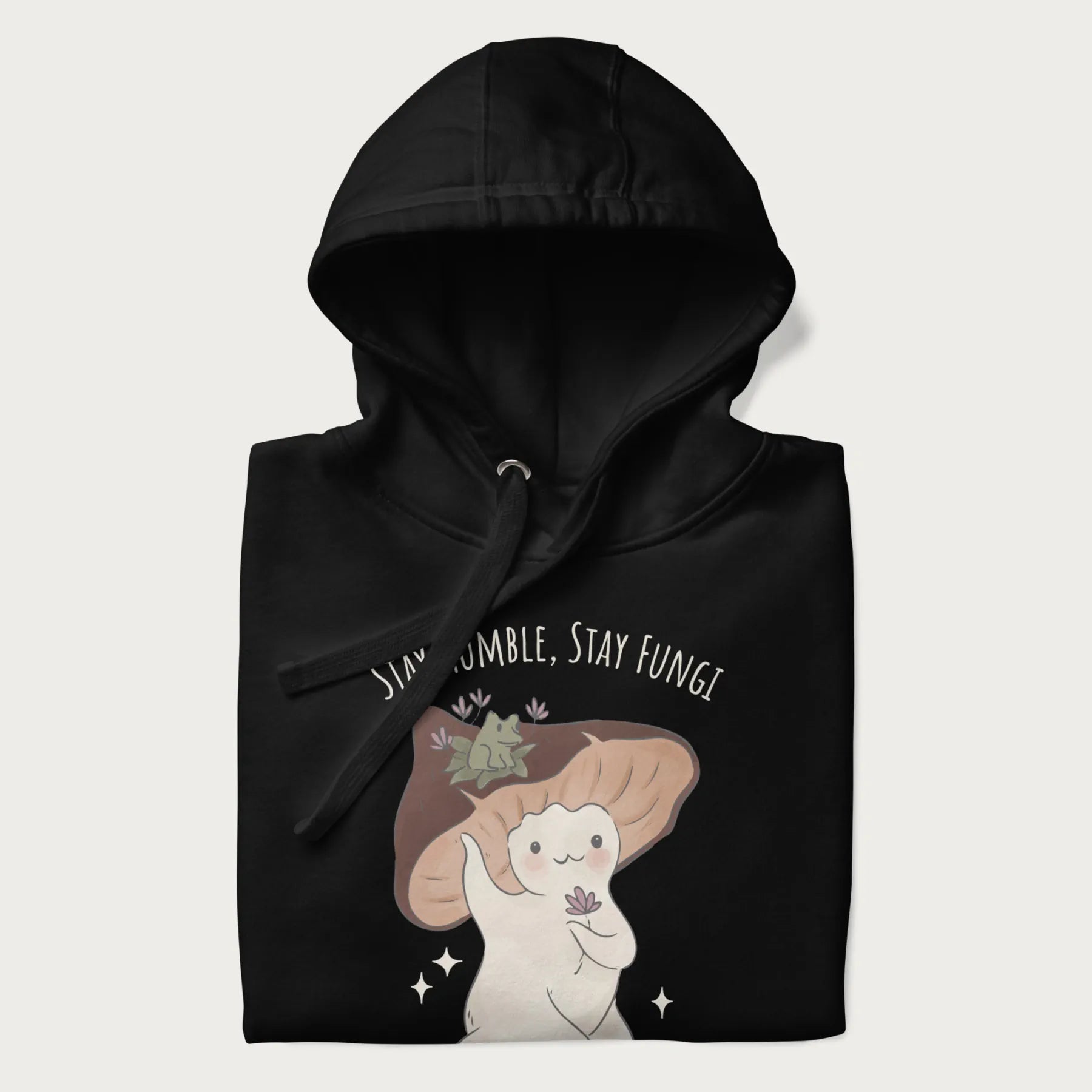 Folded black hoodie with graphic of a cute mushroom character holding a flower with a frog on its cap and the text 'Stay Humble, Stay Fungi' above.