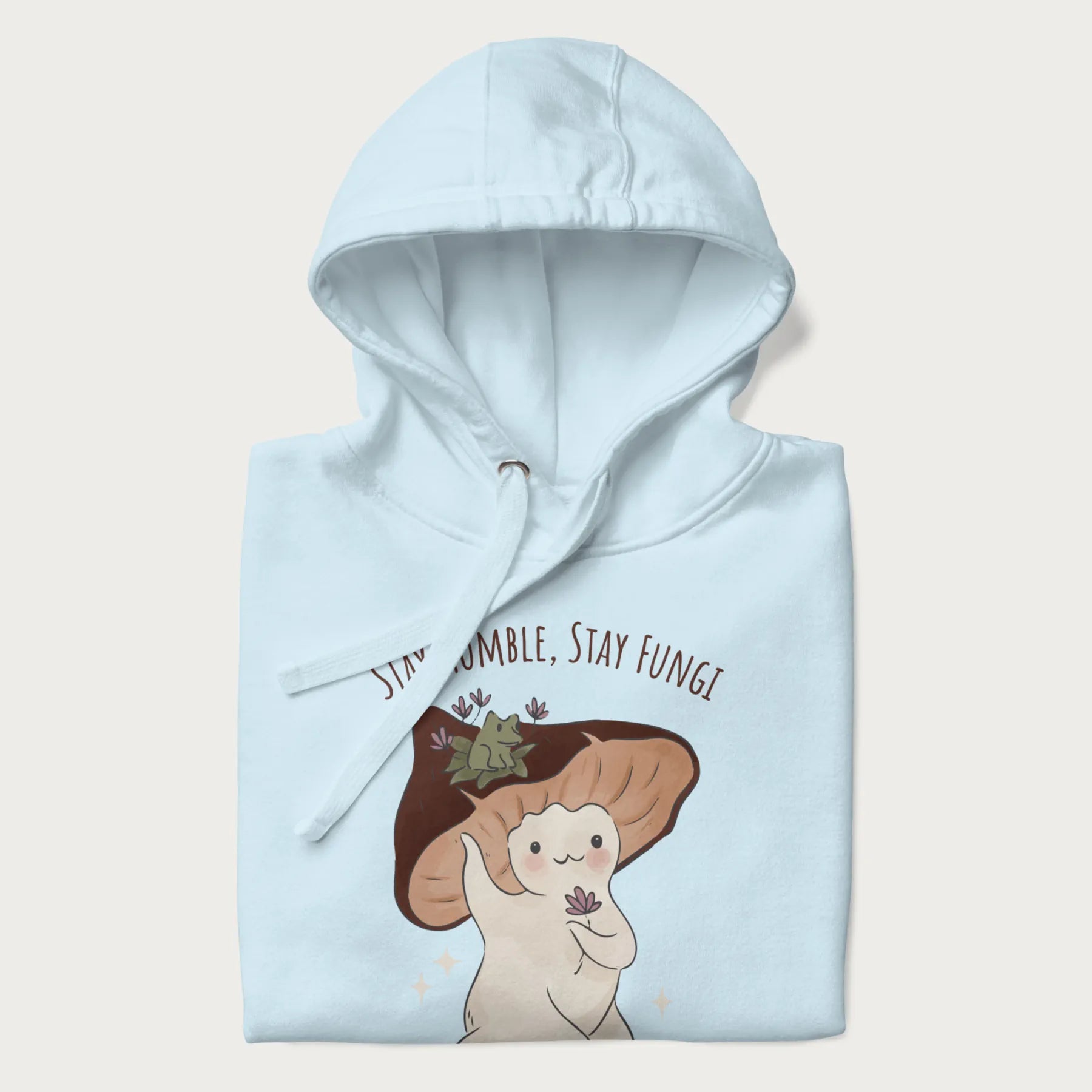 Folded light blue hoodie with graphic of a cute mushroom character holding a flower with a frog on its cap and the text 'Stay Humble, Stay Fungi' above.