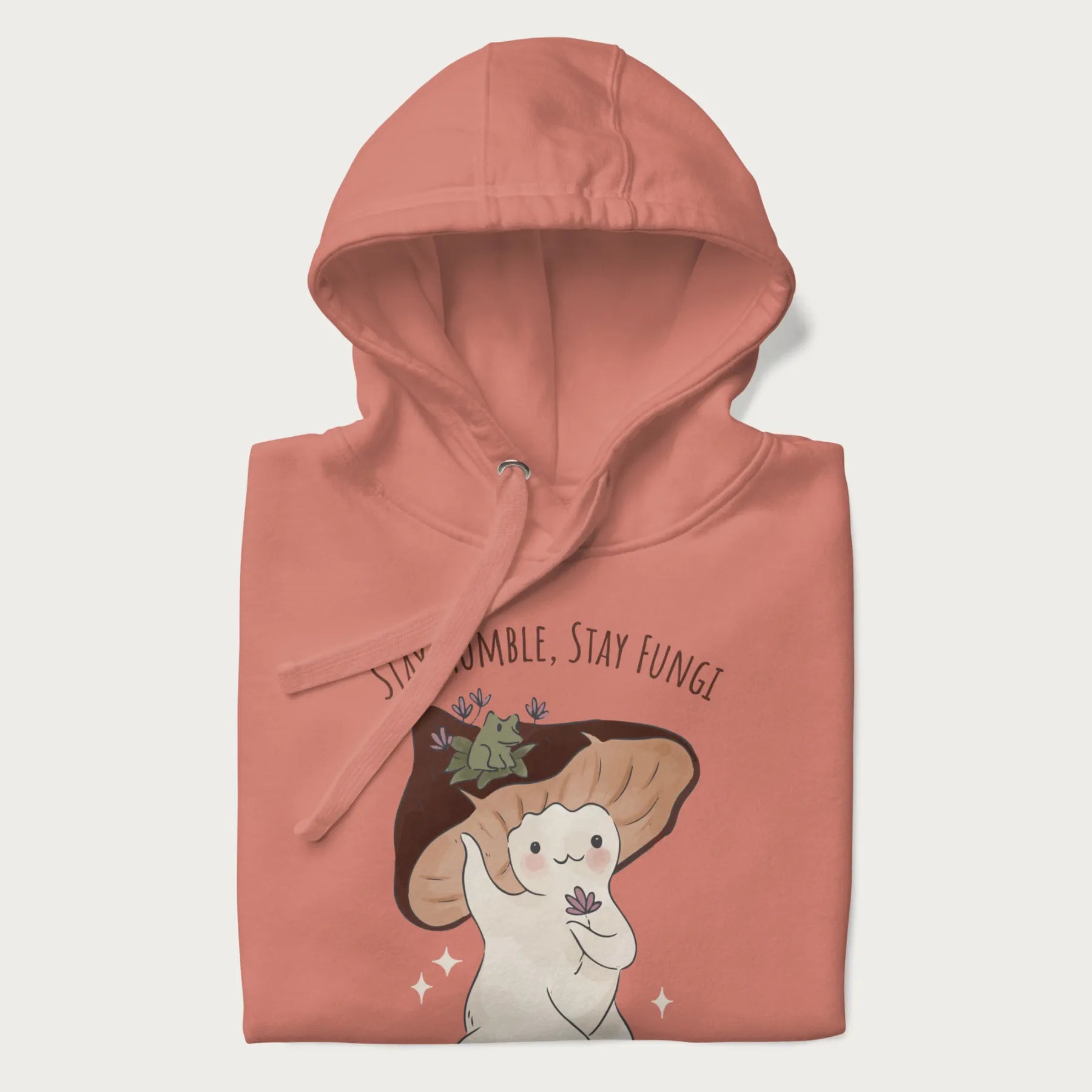 Folded light pink hoodie with graphic of a cute mushroom character holding a flower with a frog on its cap and the text 'Stay Humble, Stay Fungi' above.