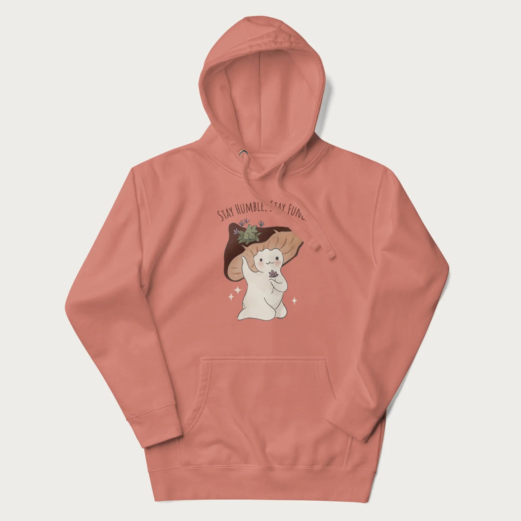 Light pink hoodie with graphic of a cute mushroom character holding a flower with a frog on its cap and the text 'Stay Humble, Stay Fungi' above.