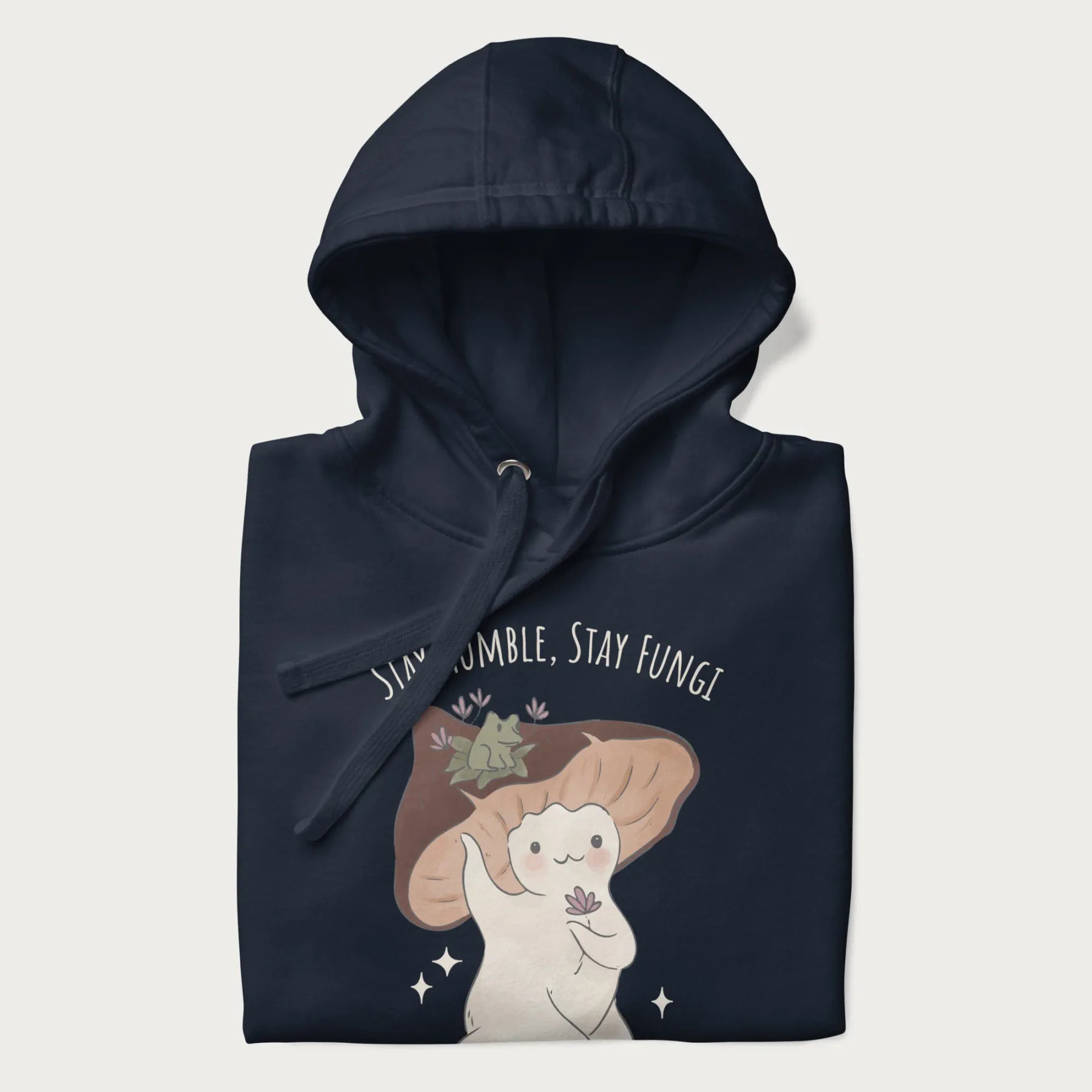 Folded navy blue hoodie with graphic of a cute mushroom character holding a flower with a frog on its cap and the text 'Stay Humble, Stay Fungi' above.