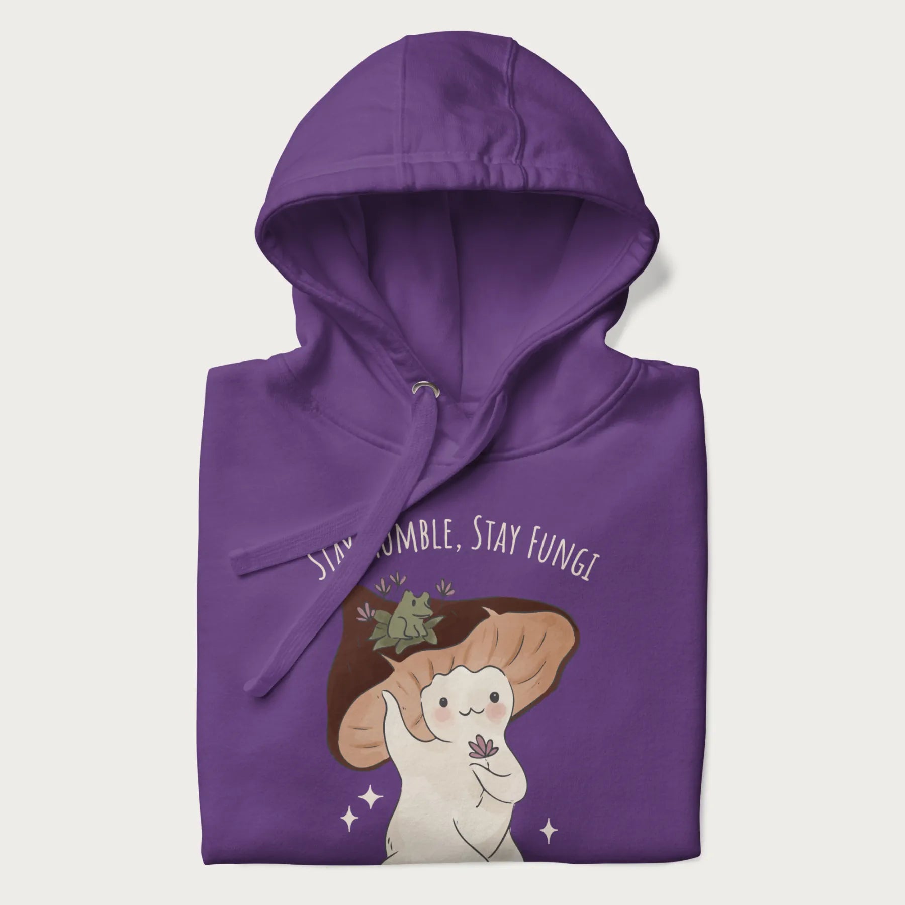 Folded purple hoodie with graphic of a cute mushroom character holding a flower with a frog on its cap and the text 'Stay Humble, Stay Fungi' above.