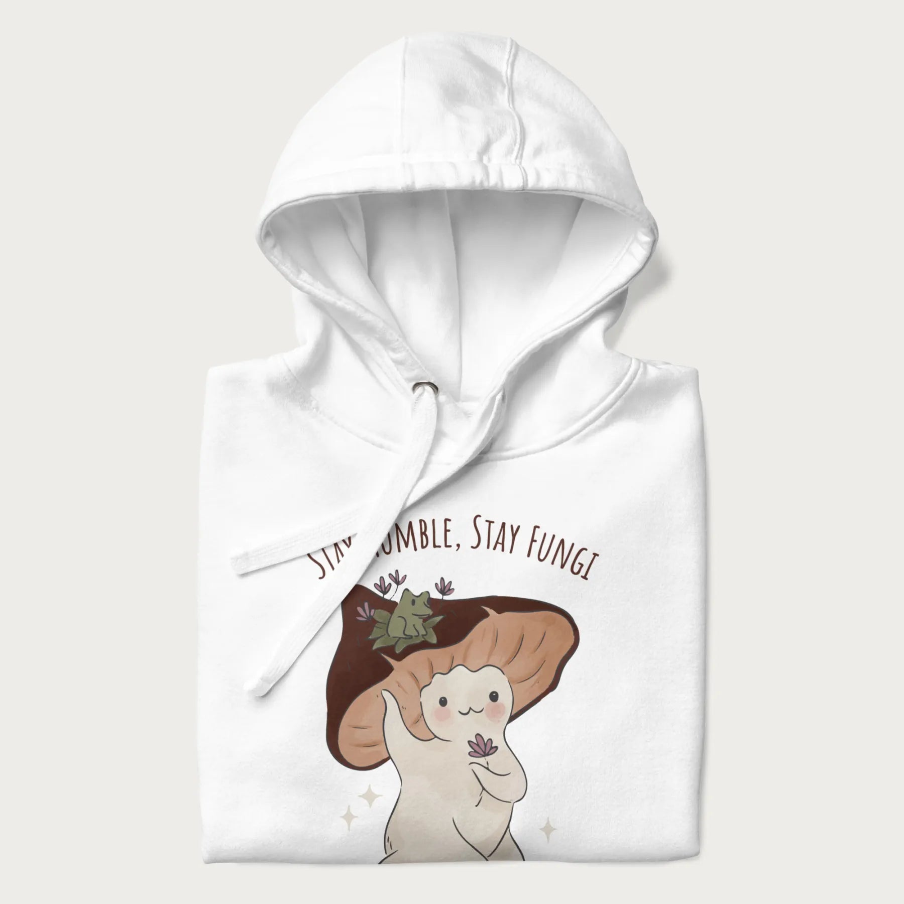 Folded white hoodie with graphic of a cute mushroom character holding a flower with a frog on its cap and the text 'Stay Humble, Stay Fungi' above.