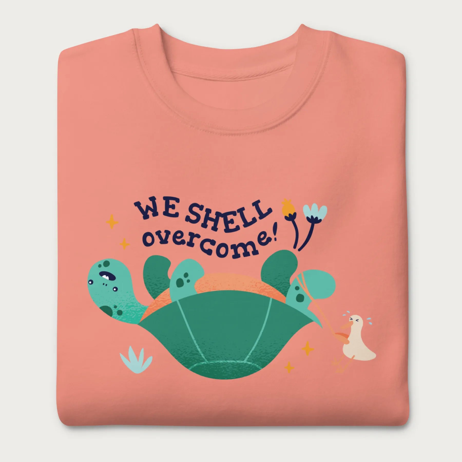 Folded light pink sweatshirt with a graphic of a turtle flipped on it's shell with the phrase 'We Shell Overcome.'