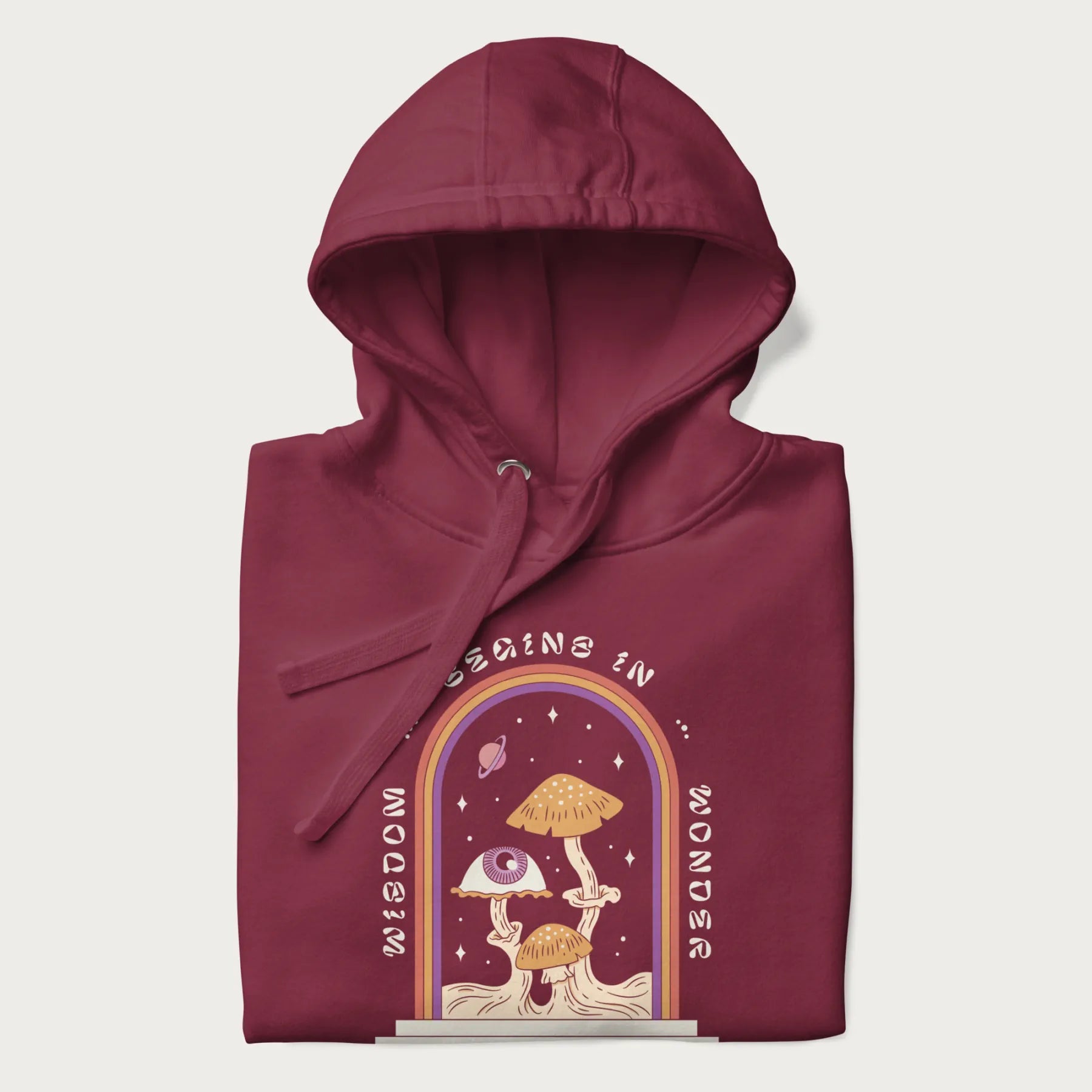 Folded maroon hoodie with graphic of mystical mushrooms with an all-seeing eye and the text 'Wisdom Begins in Wonder' framed by a rainbow arch.