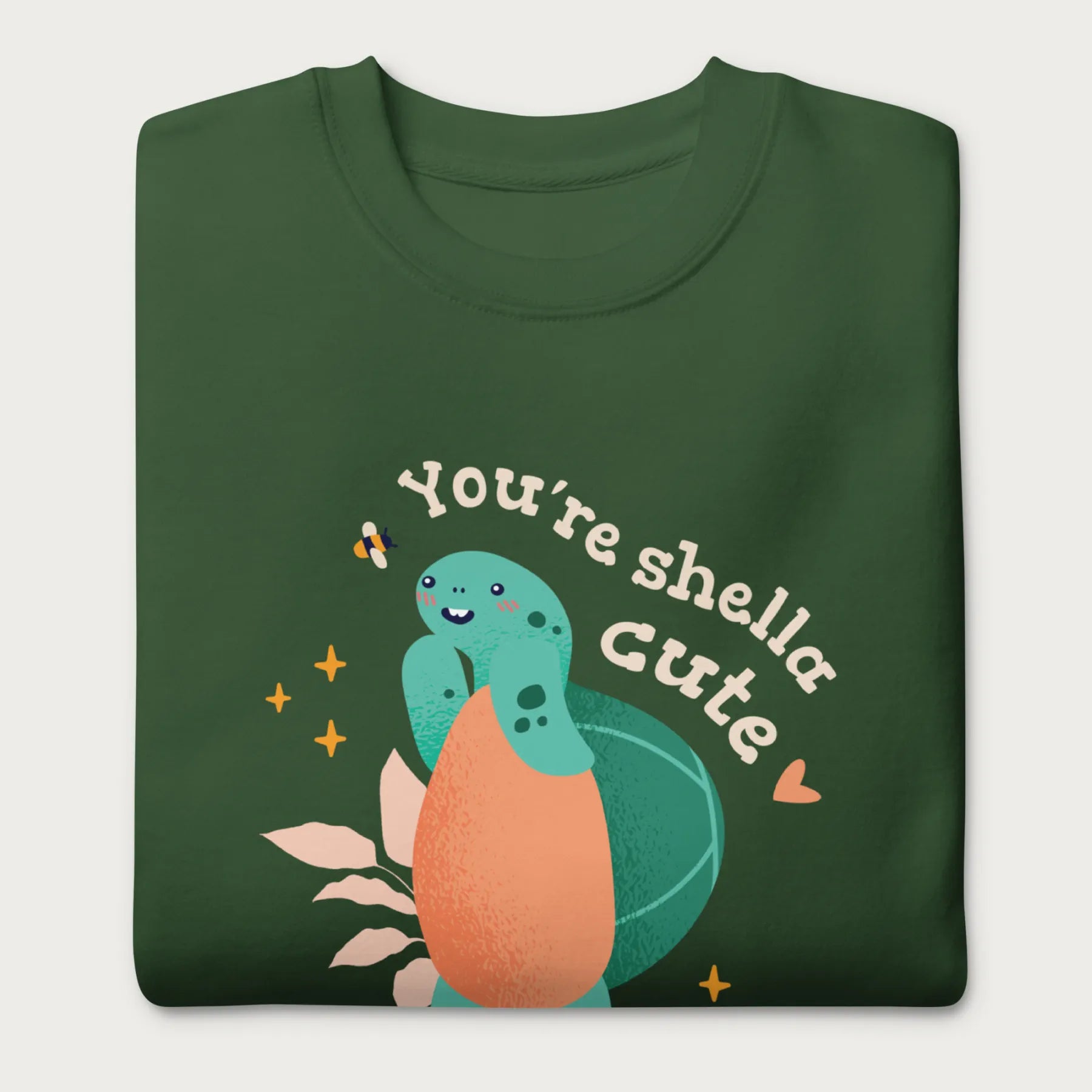 Folded forest green sweatshirt with a cute graphic of a smiling turtle and bee with the phrase 'You're Shella Cute.'
