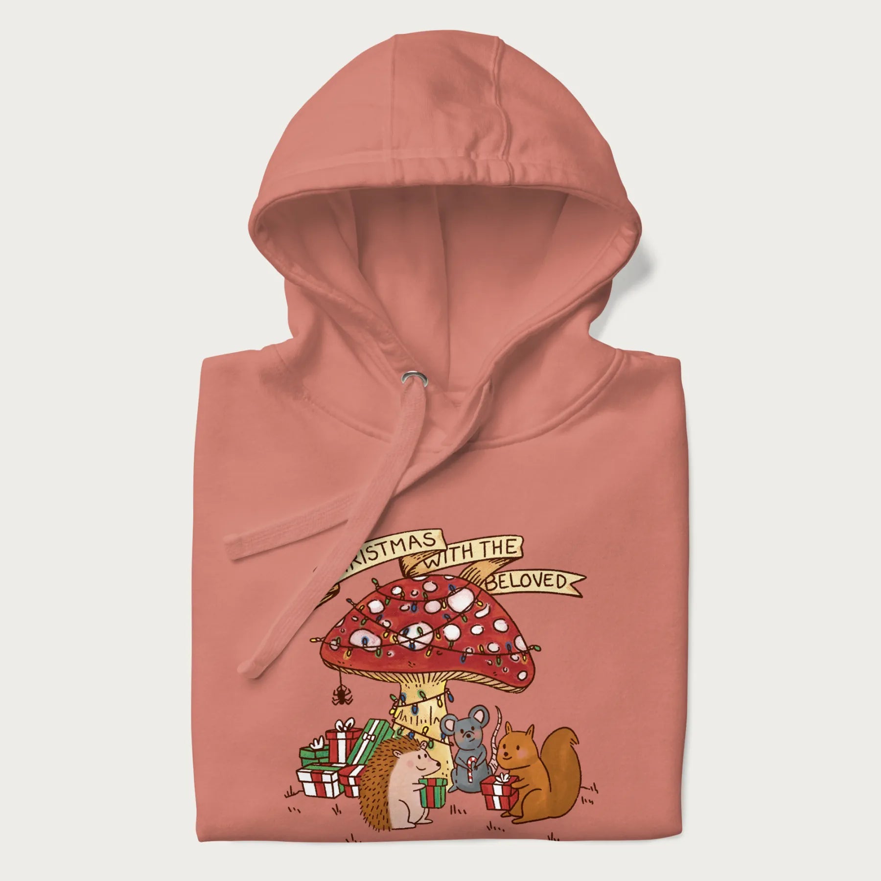 Folded light pink hoodie with animal around a festively decorated mushroom and the text 'Christmas with the Beloved' above.