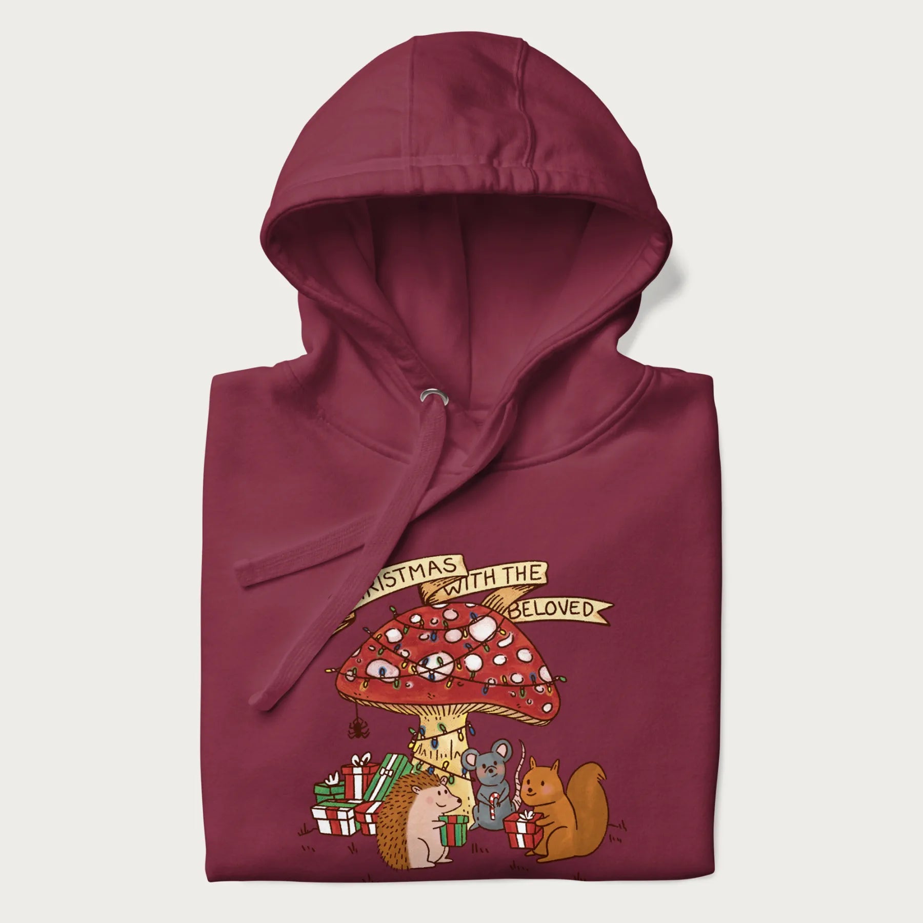 Folded maroon hoodie with animal around a festively decorated mushroom and the text 'Christmas with the Beloved' above.