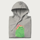 Folded light grey hoodie with a graphic of a green frog wearing a mushroom cap surrounded by stars.