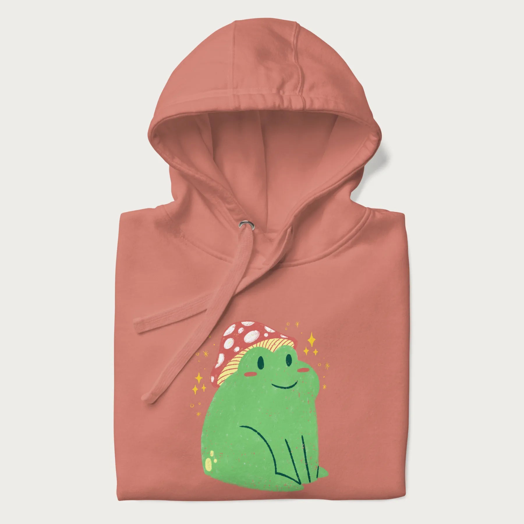 Folded light pink hoodie with a graphic of a green frog wearing a mushroom cap surrounded by stars.