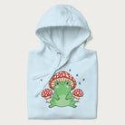 Folded light blue hoodie will illustration of a cute green frog with red and white mushrooms.