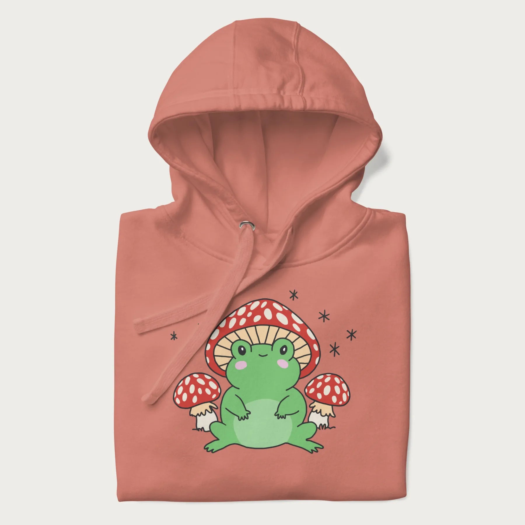 Folded light pink hoodie will illustration of a cute green frog with red and white mushrooms.