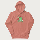 Light pink hoodie will illustration of a cute green frog with red and white mushrooms.