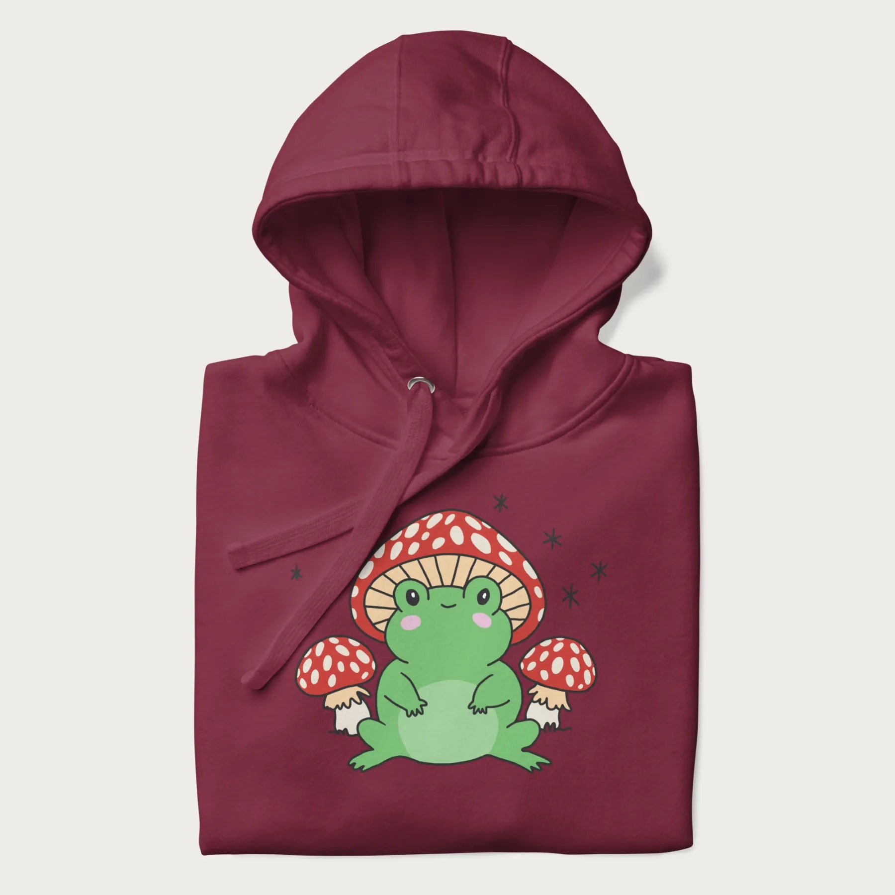 Folded maroon hoodie will illustration of a cute green frog with red and white mushrooms.