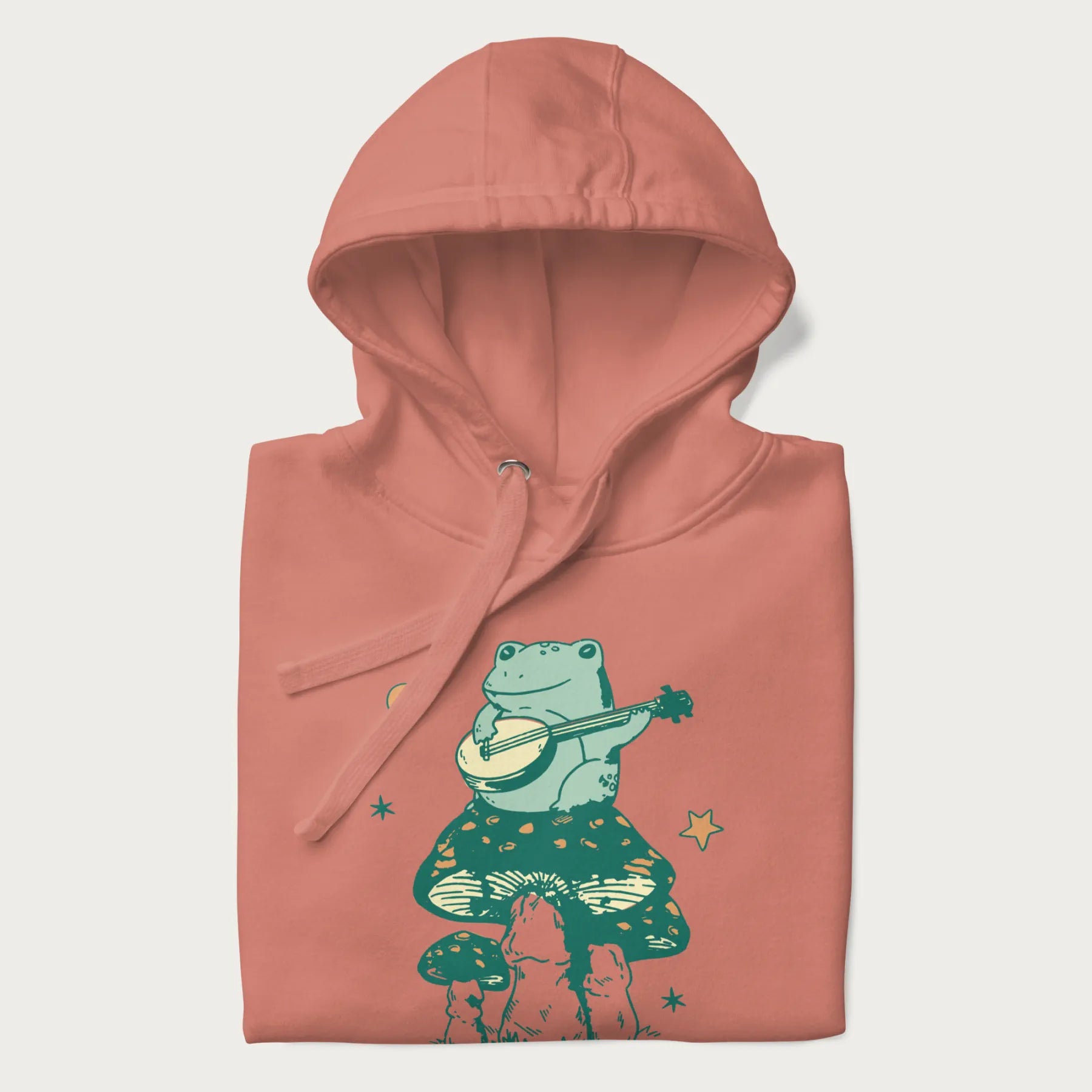 Folded light pink hoodie with graphic of a frog playing a banjo on a mushroom with stars and a crescent moon.