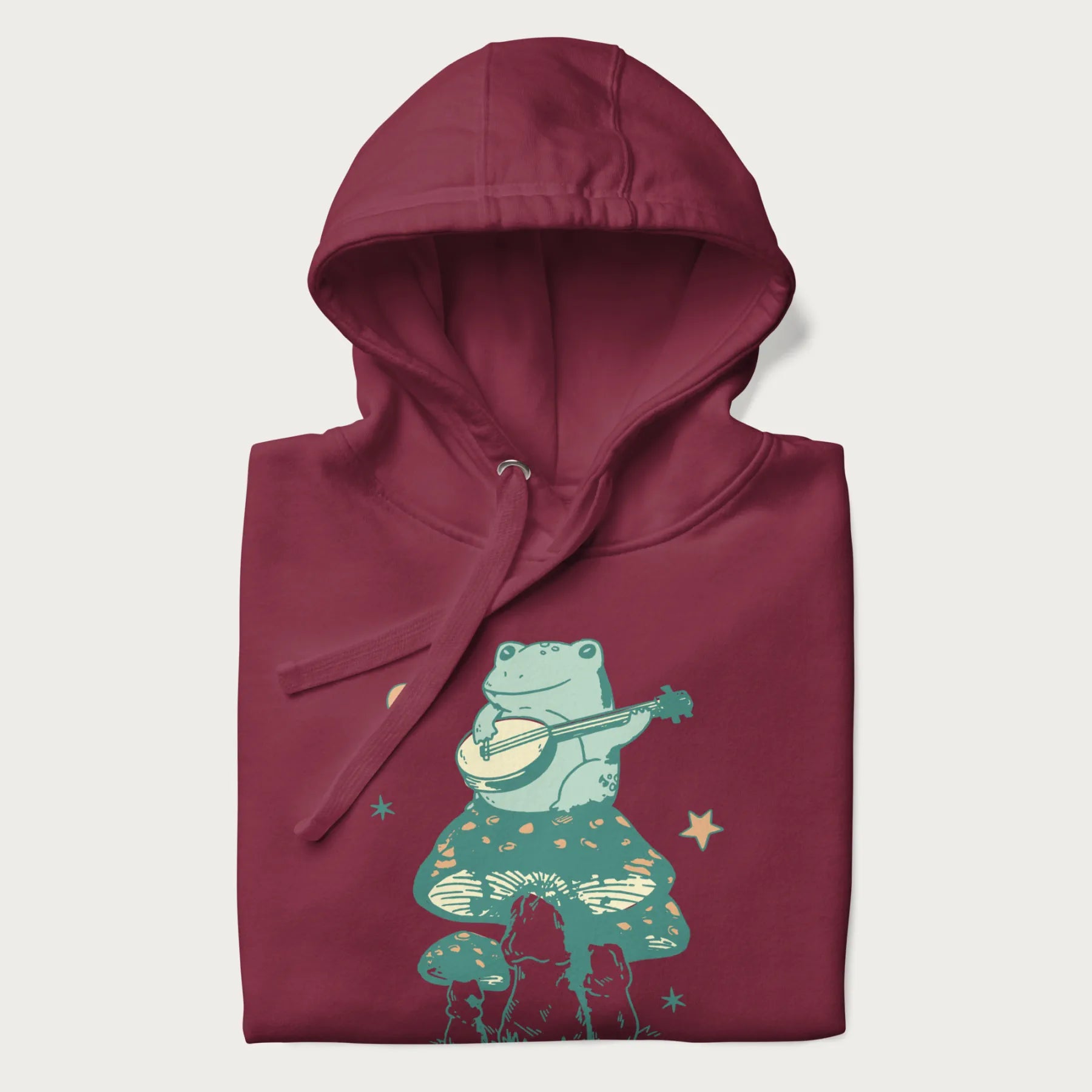 Folded maroon hoodie with graphic of a frog playing a banjo on a mushroom with stars and a crescent moon.