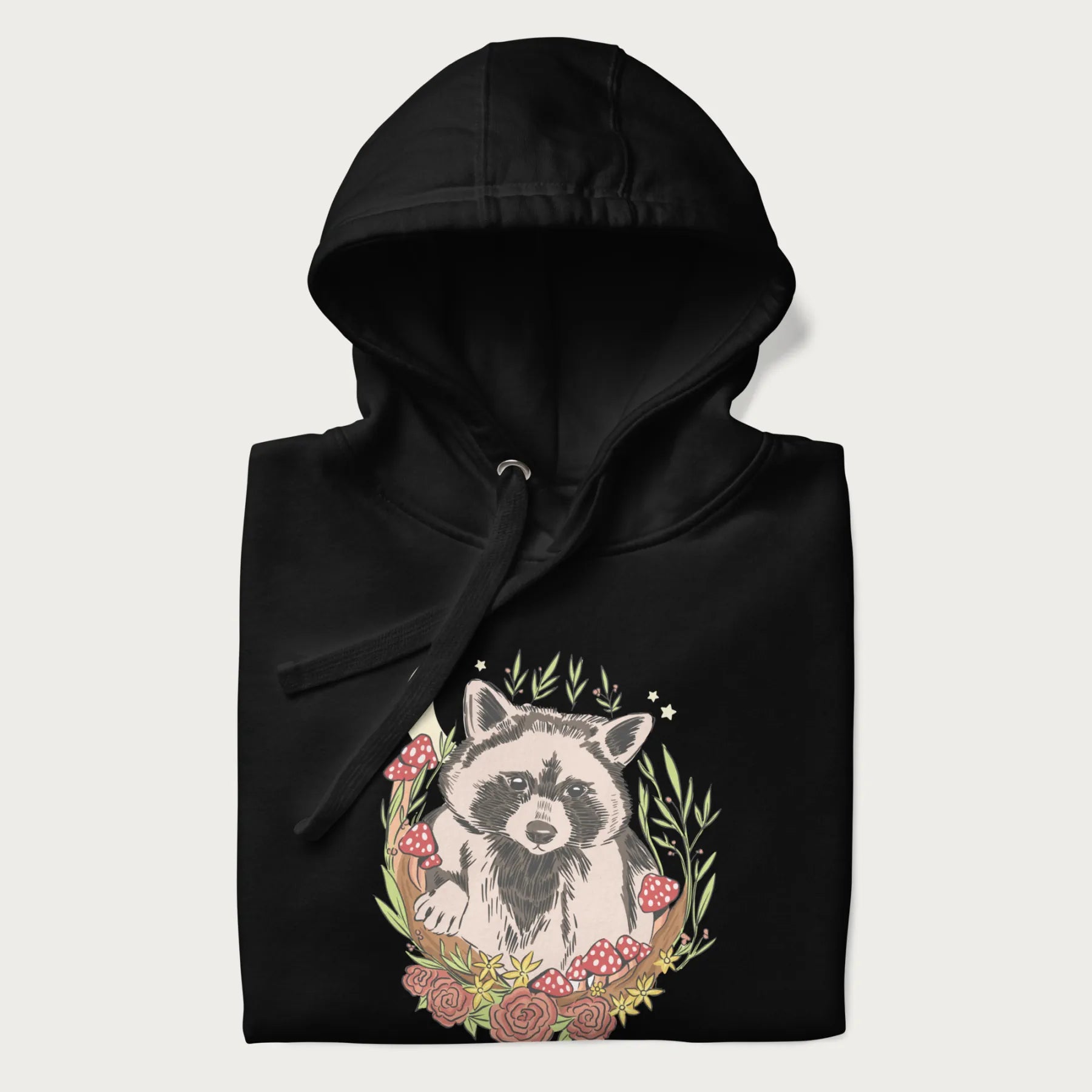 Folded black hoodie with graphic of an adorable raccoon surrounded by mushrooms, foliage, and a crescent moon with stars in the background.