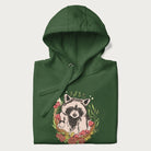 Folded forest green hoodie with graphic of an adorable raccoon surrounded by mushrooms, foliage, and a crescent moon with stars in the background.