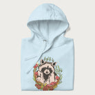 Folded light blue hoodie with graphic of an adorable raccoon surrounded by mushrooms, foliage, and a crescent moon with stars in the background.