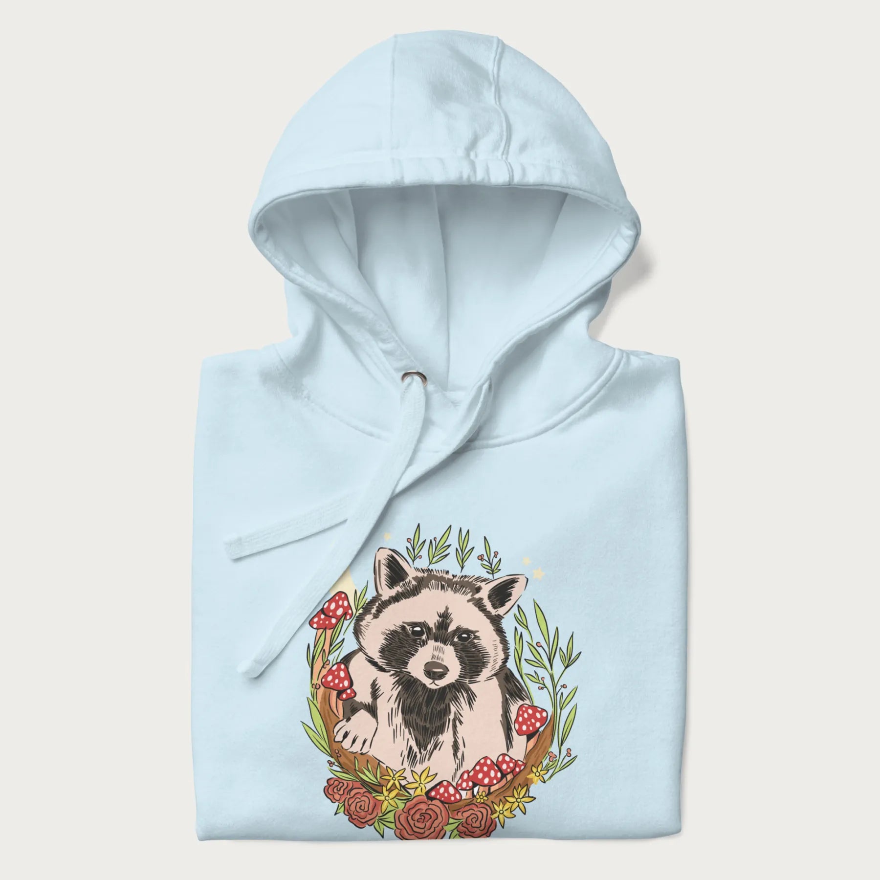 Folded light blue hoodie with graphic of an adorable raccoon surrounded by mushrooms, foliage, and a crescent moon with stars in the background.