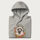 Folded light grey hoodie with graphic of an adorable raccoon surrounded by mushrooms, foliage, and a crescent moon with stars in the background.