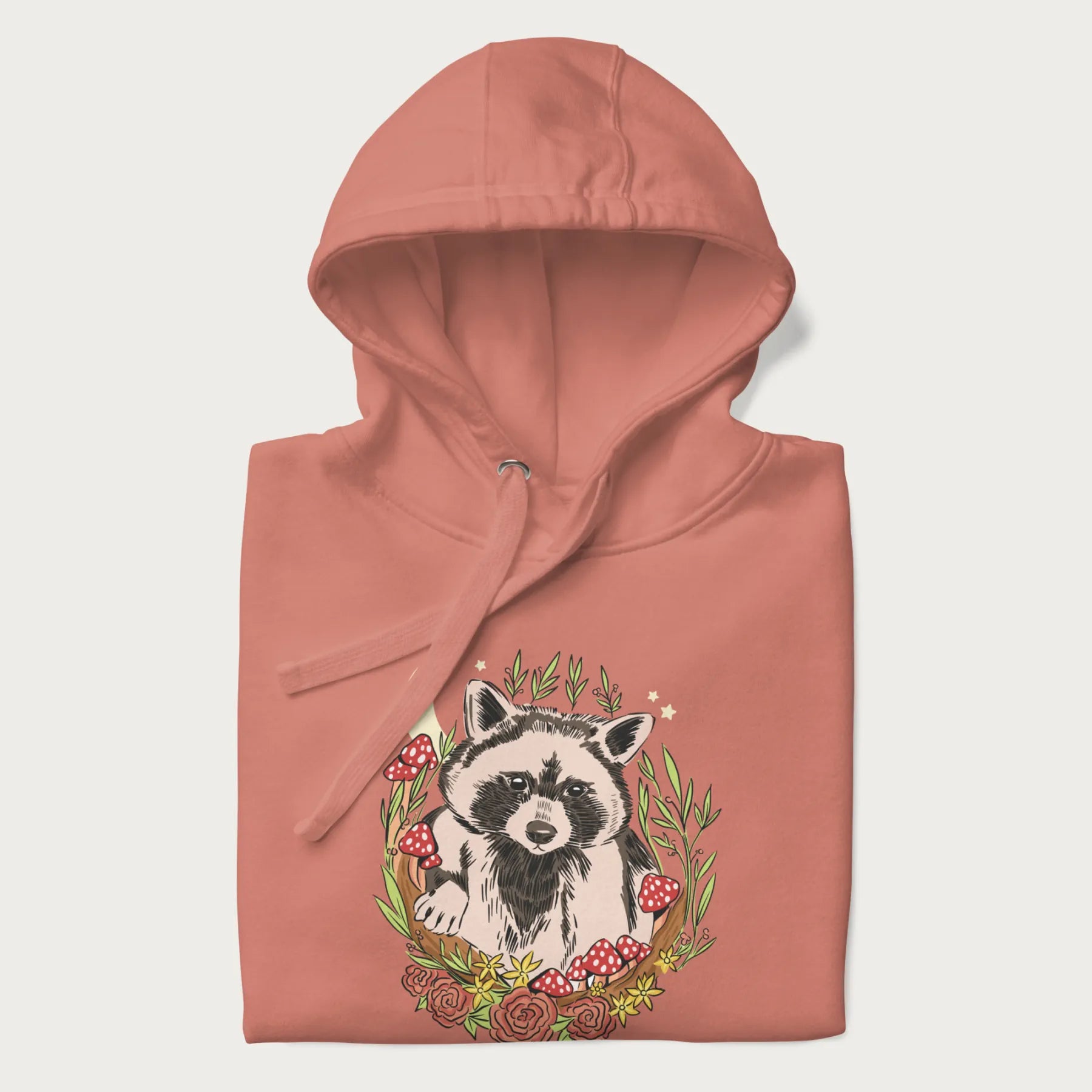 Folded light pink hoodie with graphic of an adorable raccoon surrounded by mushrooms, foliage, and a crescent moon with stars in the background.