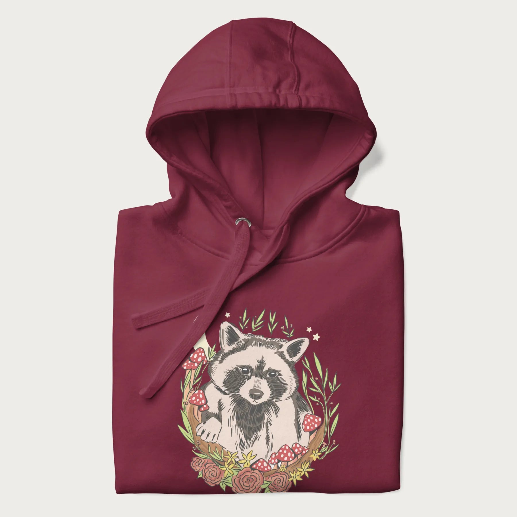 Folded maroon hoodie with graphic of an adorable raccoon surrounded by mushrooms, foliage, and a crescent moon with stars in the background.