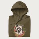 Folded military green hoodie with graphic of an adorable raccoon surrounded by mushrooms, foliage, and a crescent moon with stars in the background.