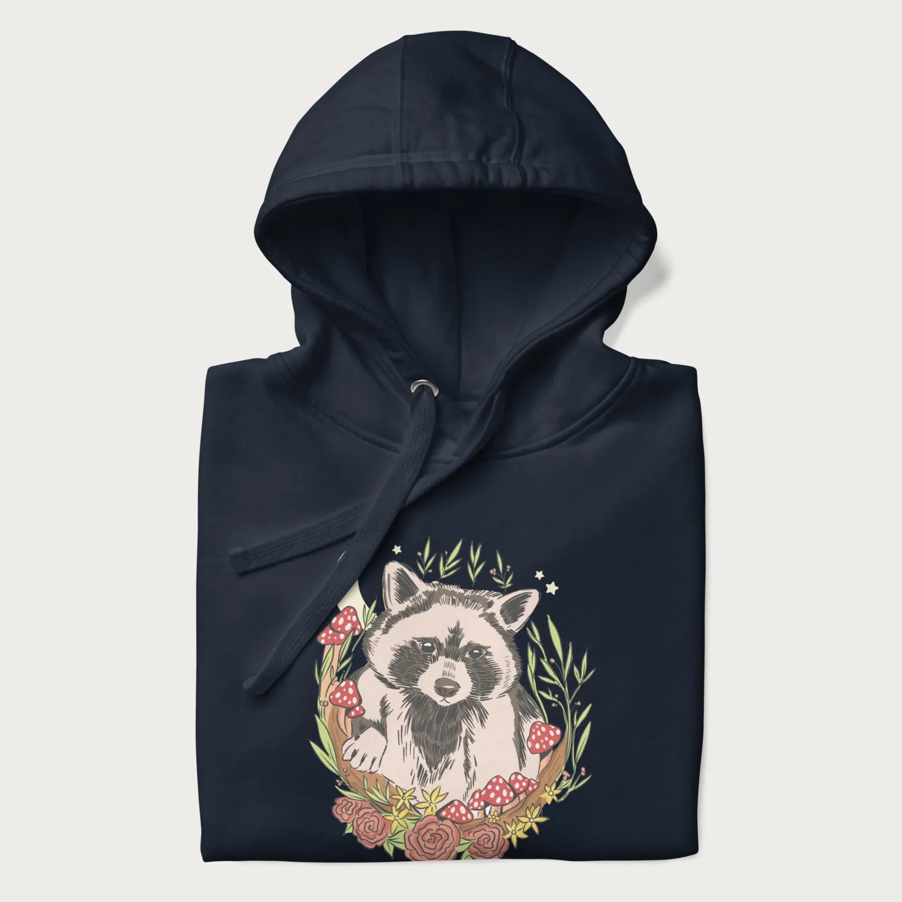 Folded navy blue hoodie with graphic of an adorable raccoon surrounded by mushrooms, foliage, and a crescent moon with stars in the background.