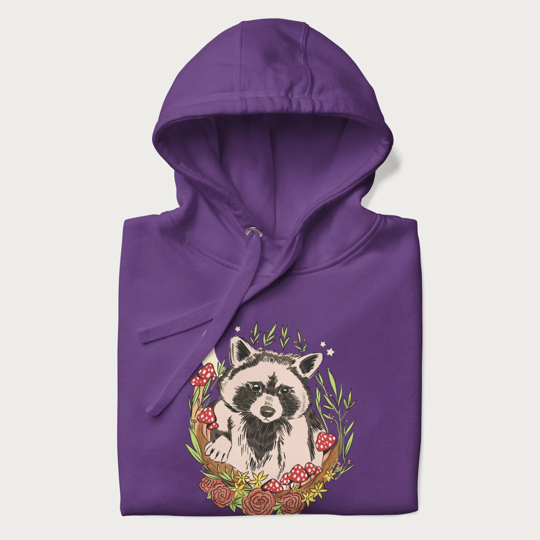 Folded purple hoodie with graphic of an adorable raccoon surrounded by mushrooms, foliage, and a crescent moon with stars in the background.