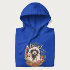 Folded royal blue hoodie with graphic of an adorable raccoon surrounded by mushrooms, foliage, and a crescent moon with stars in the background.