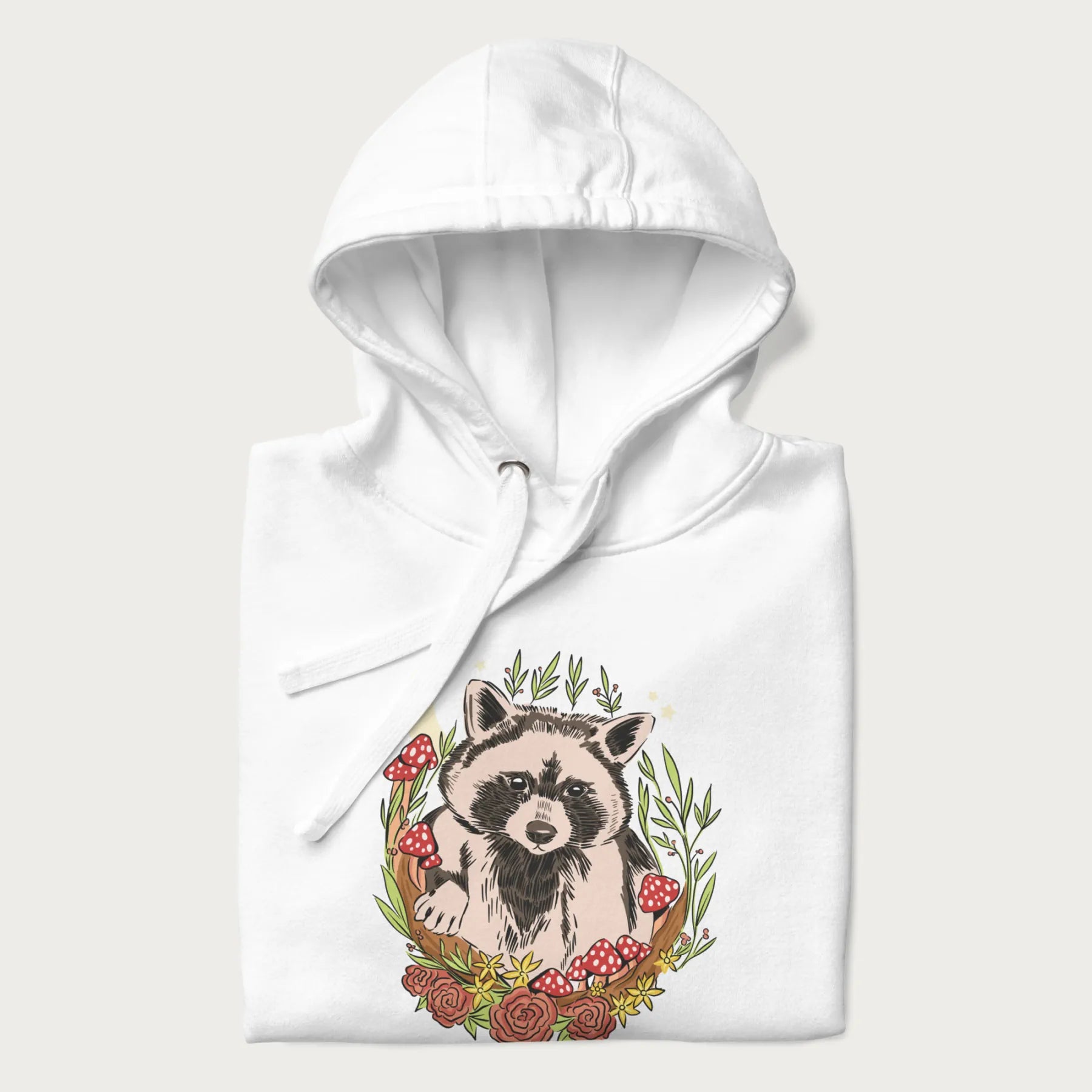 Folded white hoodie with graphic of an adorable raccoon surrounded by mushrooms, foliage, and a crescent moon with stars in the background.