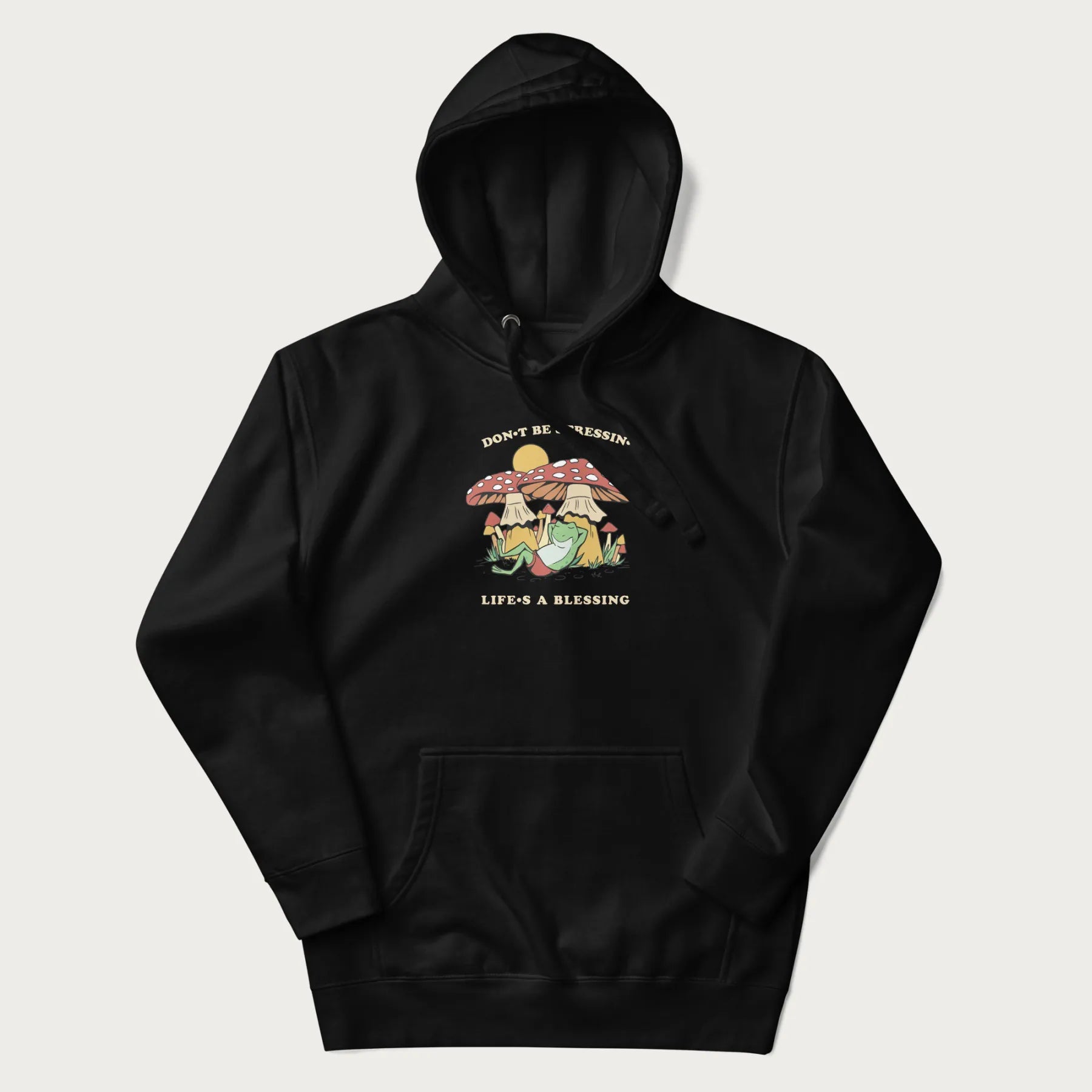 Black hoodie with a graphic of a frog lounging under mushrooms and the phrases 'Don't Be Stressin' Life's a Blessing'.
