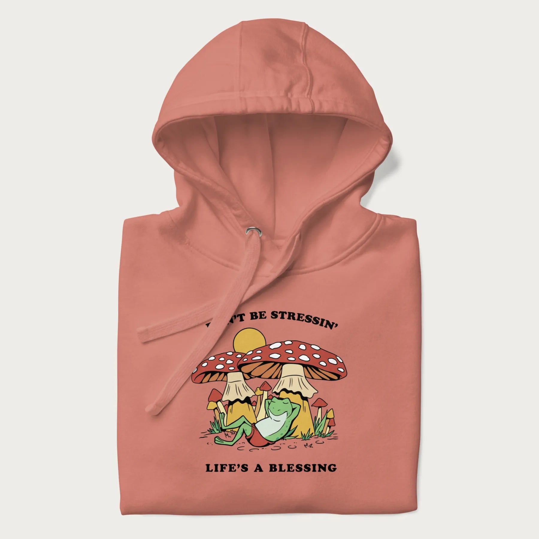 Folded light pink hoodie with a graphic of a frog lounging under mushrooms and the phrases 'Don't Be Stressin' Life's a Blessing'.