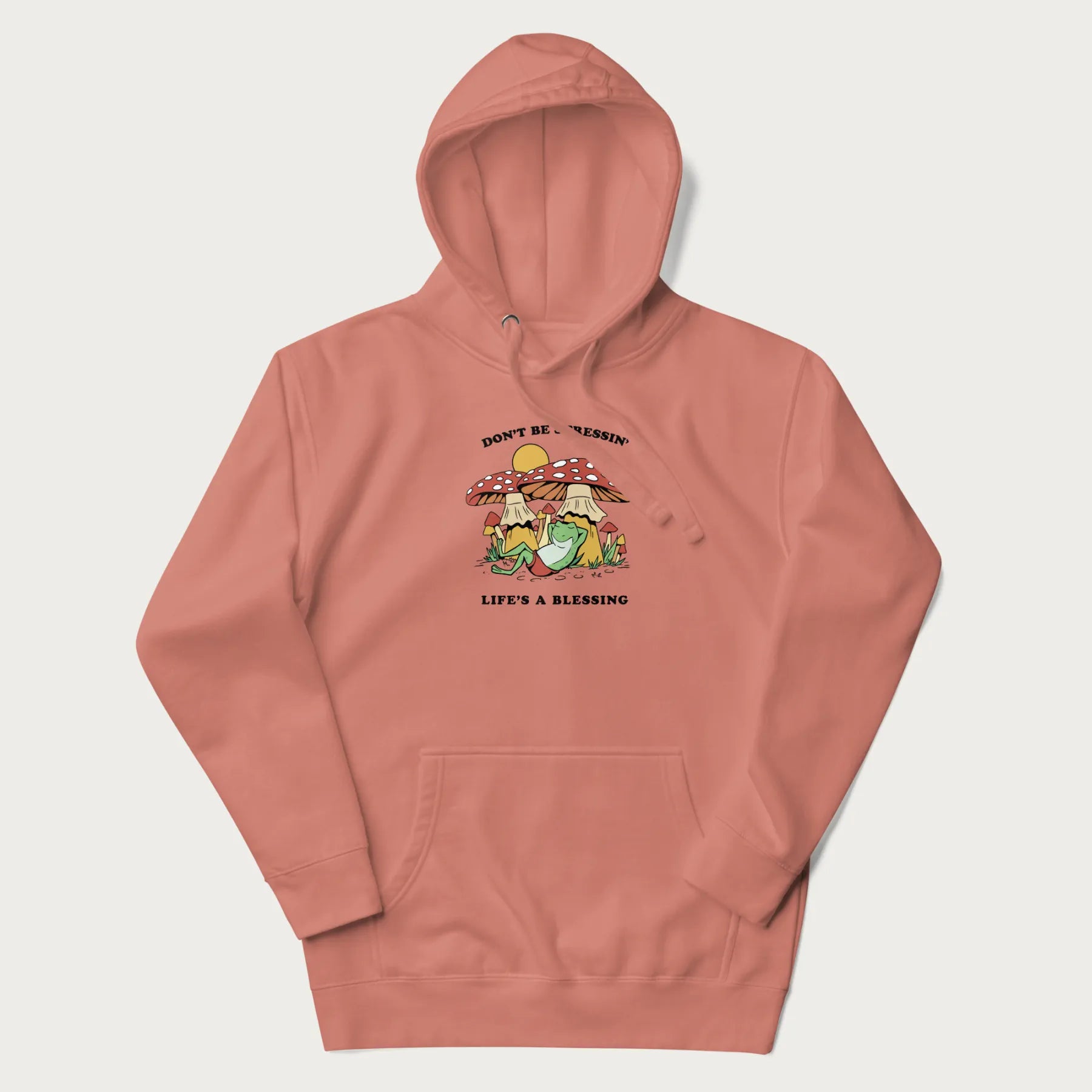 Light pink hoodie with a graphic of a frog lounging under mushrooms and the phrases 'Don't Be Stressin' Life's a Blessing'.