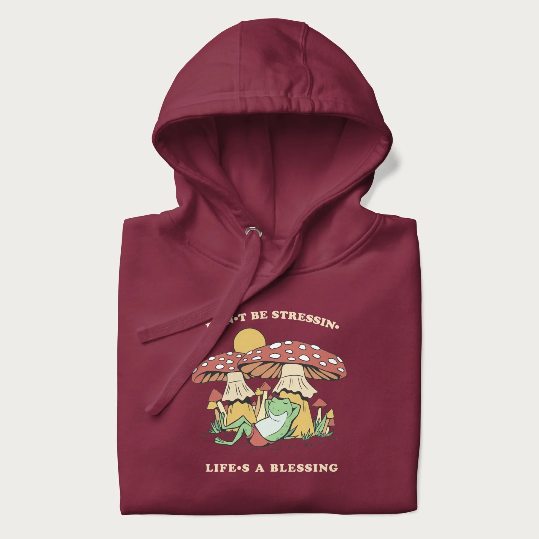 Folded maroon hoodie with a graphic of a frog lounging under mushrooms and the phrases 'Don't Be Stressin' Life's a Blessing'.