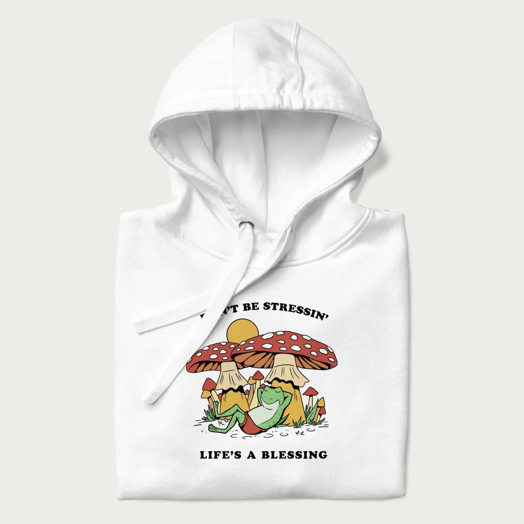 Folded white hoodie with a graphic of a frog lounging under mushrooms and the phrases 'Don't Be Stressin' Life's a Blessing'.