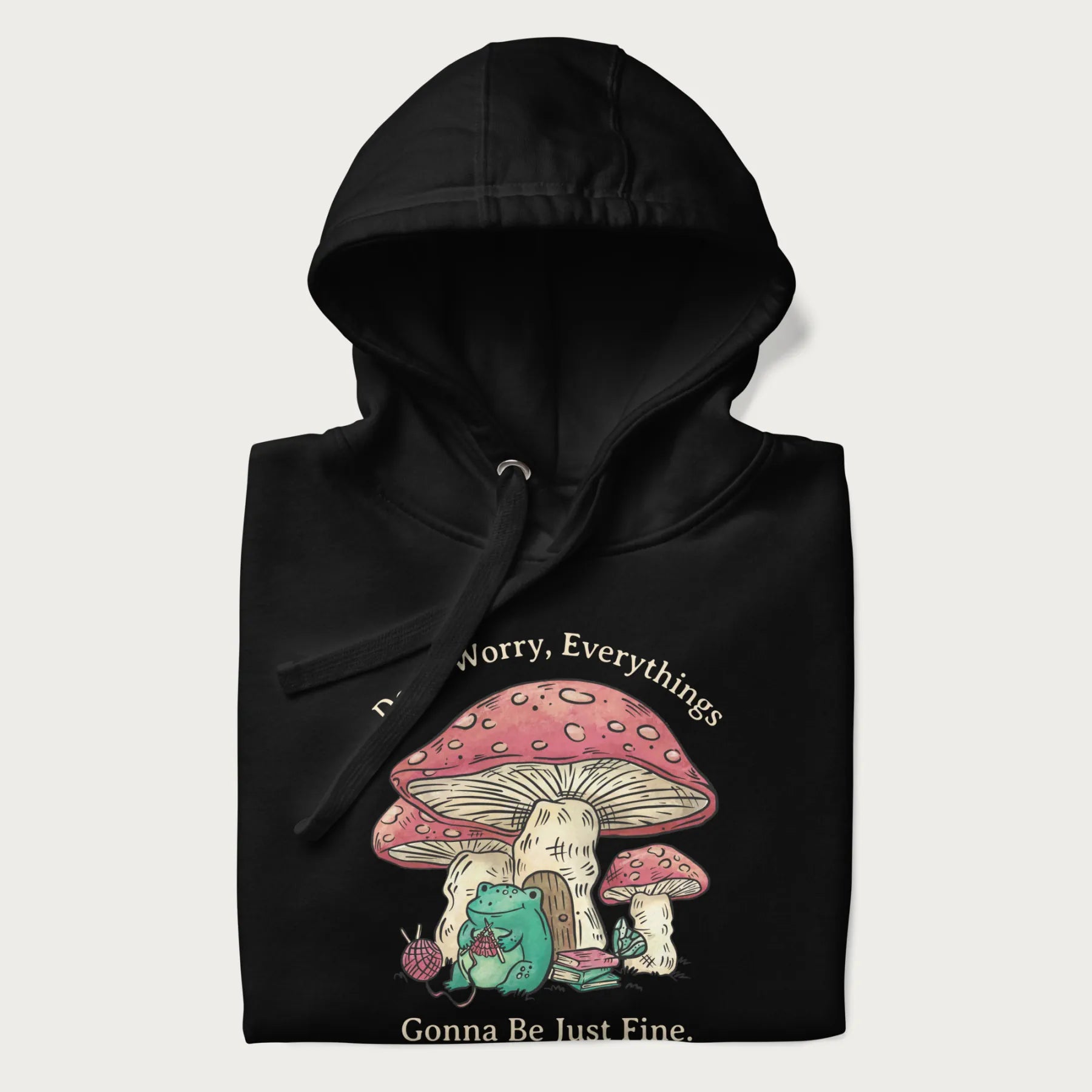 Folded black hoodie with a graphic of a frog knitting under a large mushroom with the text 'Don't Worry, Everything's Gonna Be Just Fine'.